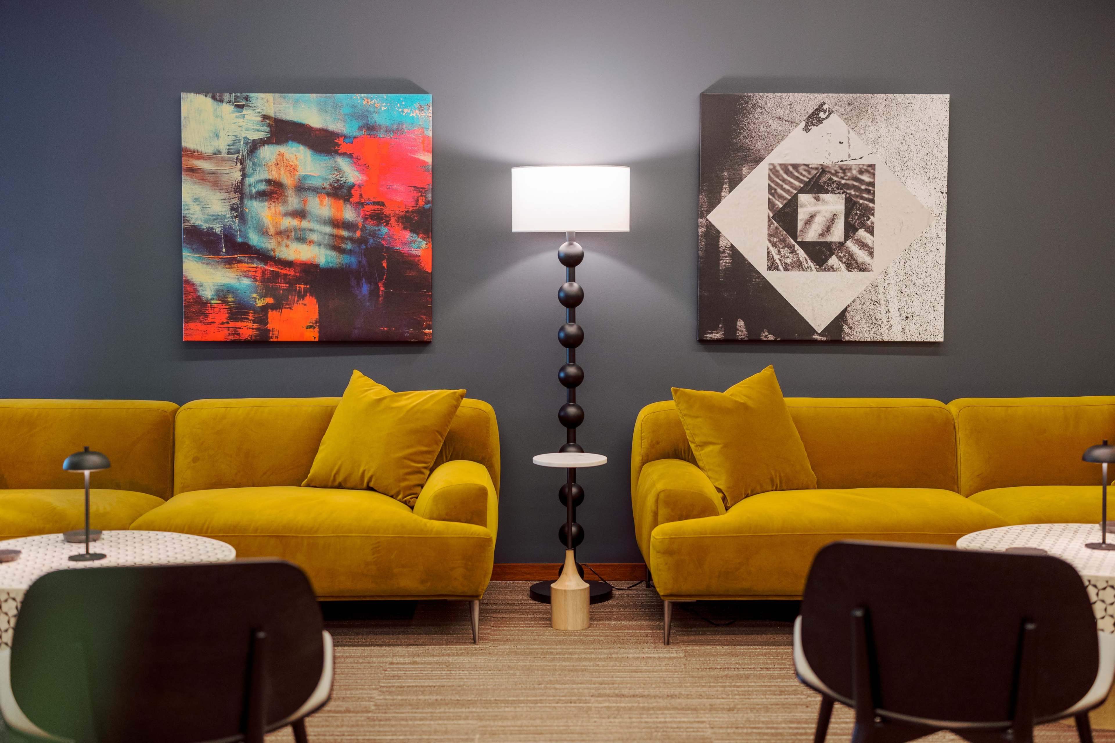 The image features a modern seating area with two yellow sofas facing each other, complemented by abstract art on the walls and a standing lamp in the center.