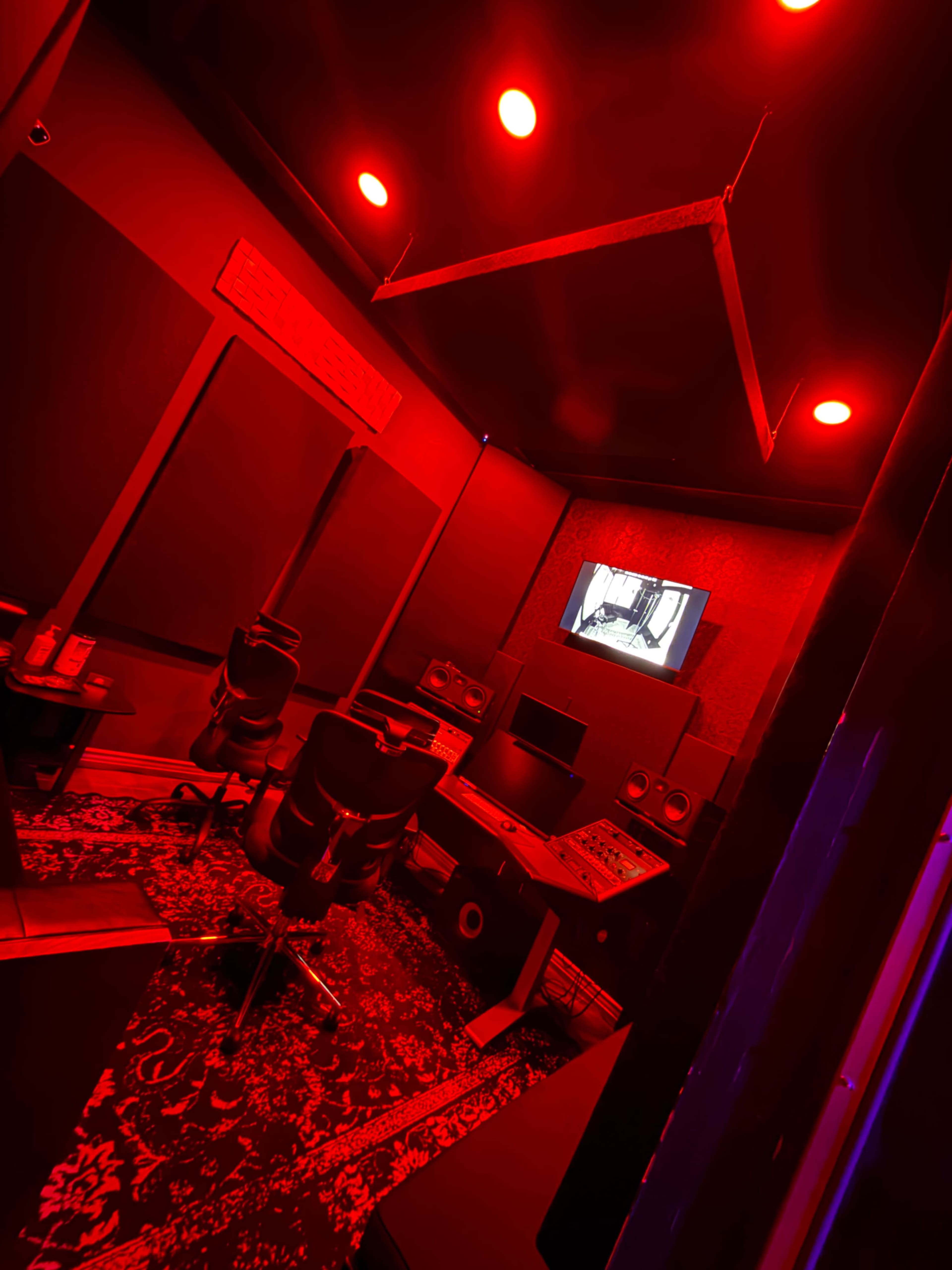 The image shows a dimly lit sound studio with red lighting, featuring a control desk, monitors, and several chairs arranged in the space.