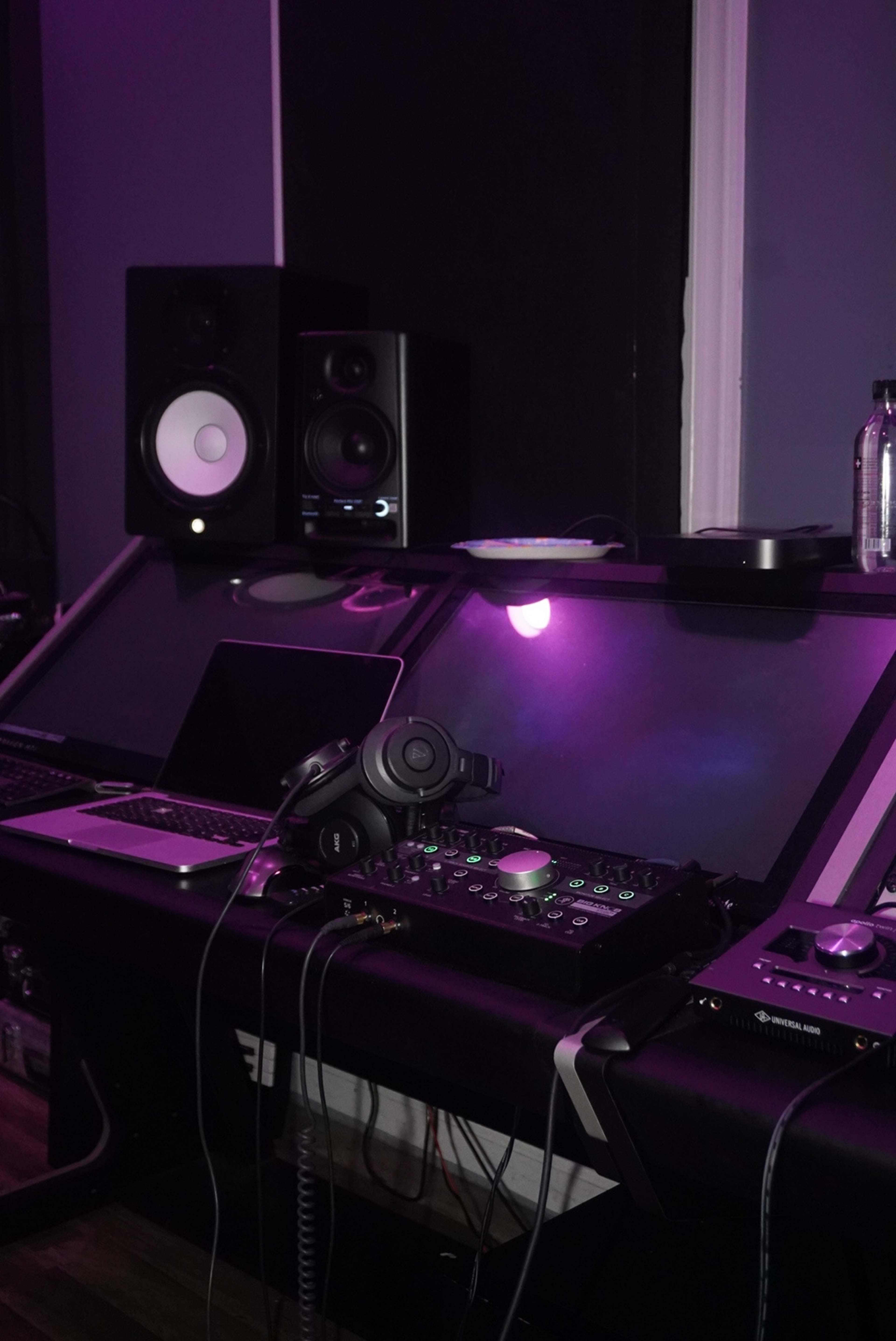 A music production setup with two speakers, a laptop, and various audio equipment illuminated by purple lighting.