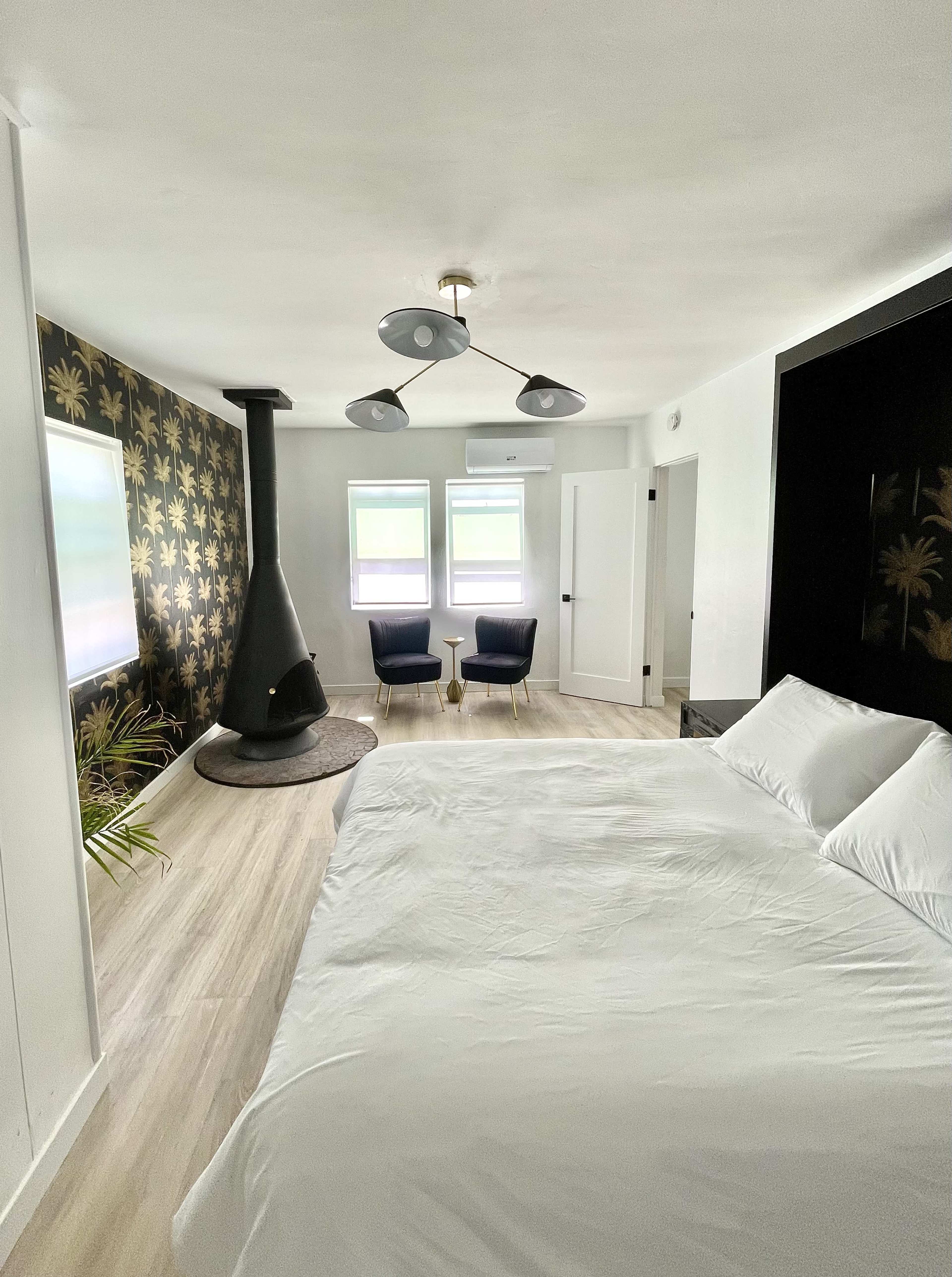 The room features a bed, two chairs, a modern light fixture, and a black fireplace against a patterned wall.