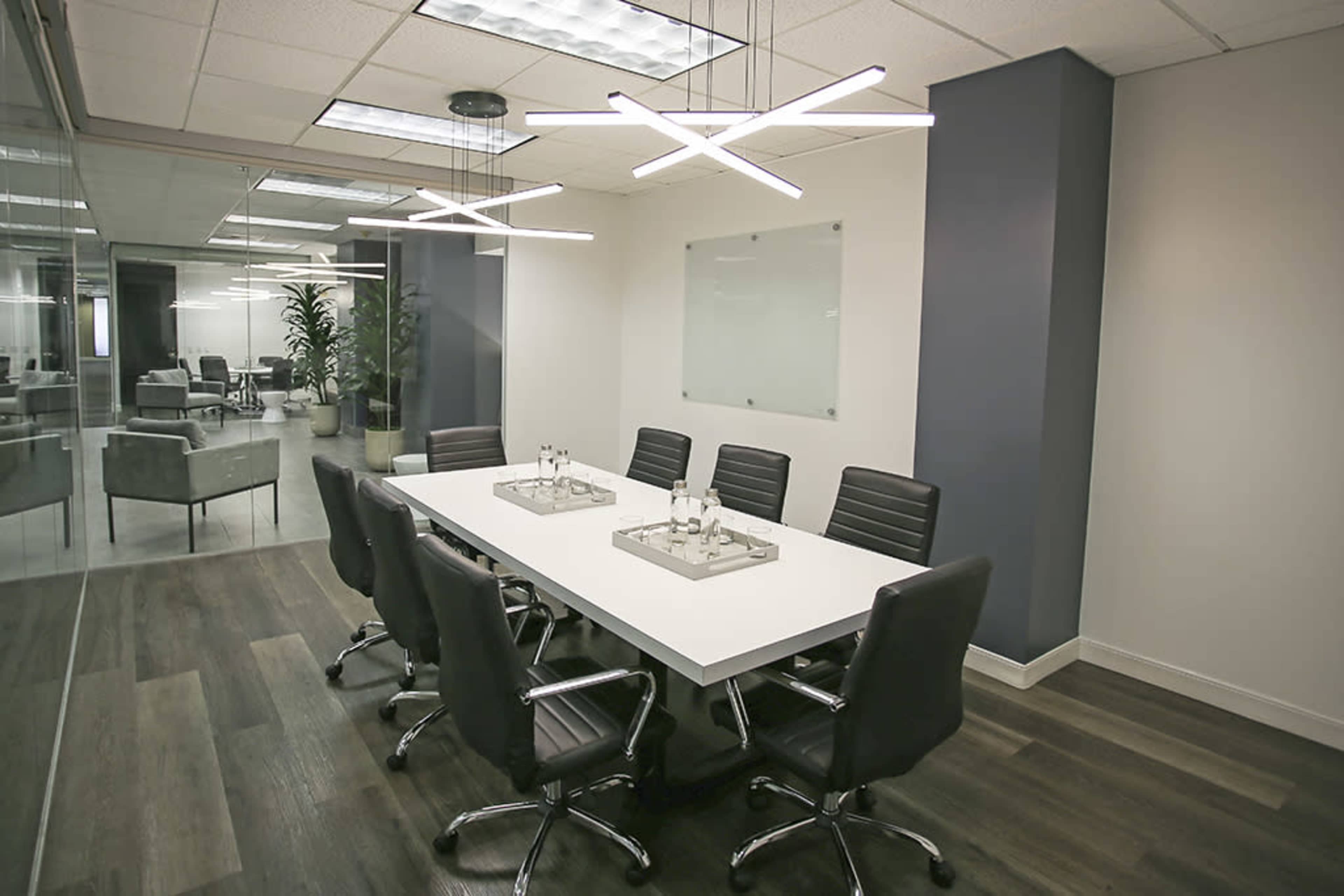 A modern conference room features a long white table surrounded by black leather chairs, with minimalistic lighting and a glass wall.