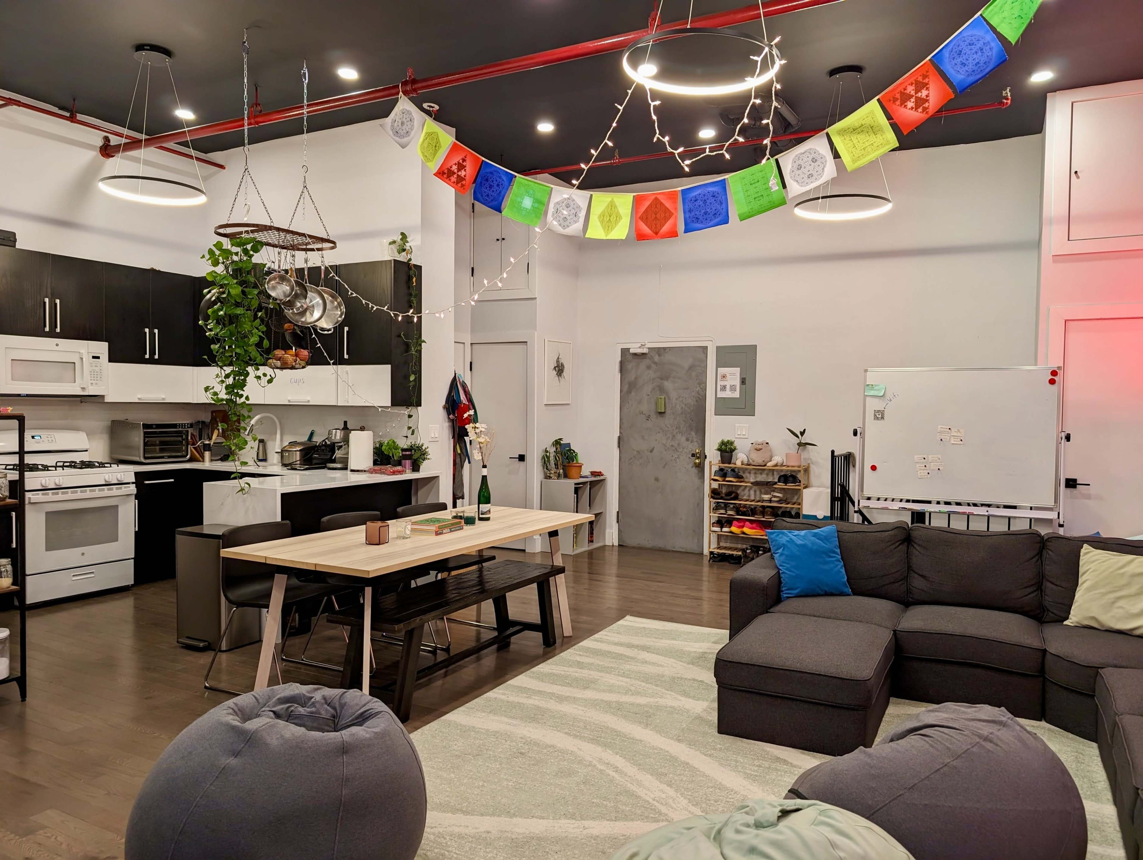 A spacious living area features a kitchen with black cabinets, a large dining table, a gray sectional couch, and colorful decorative banners overhead.