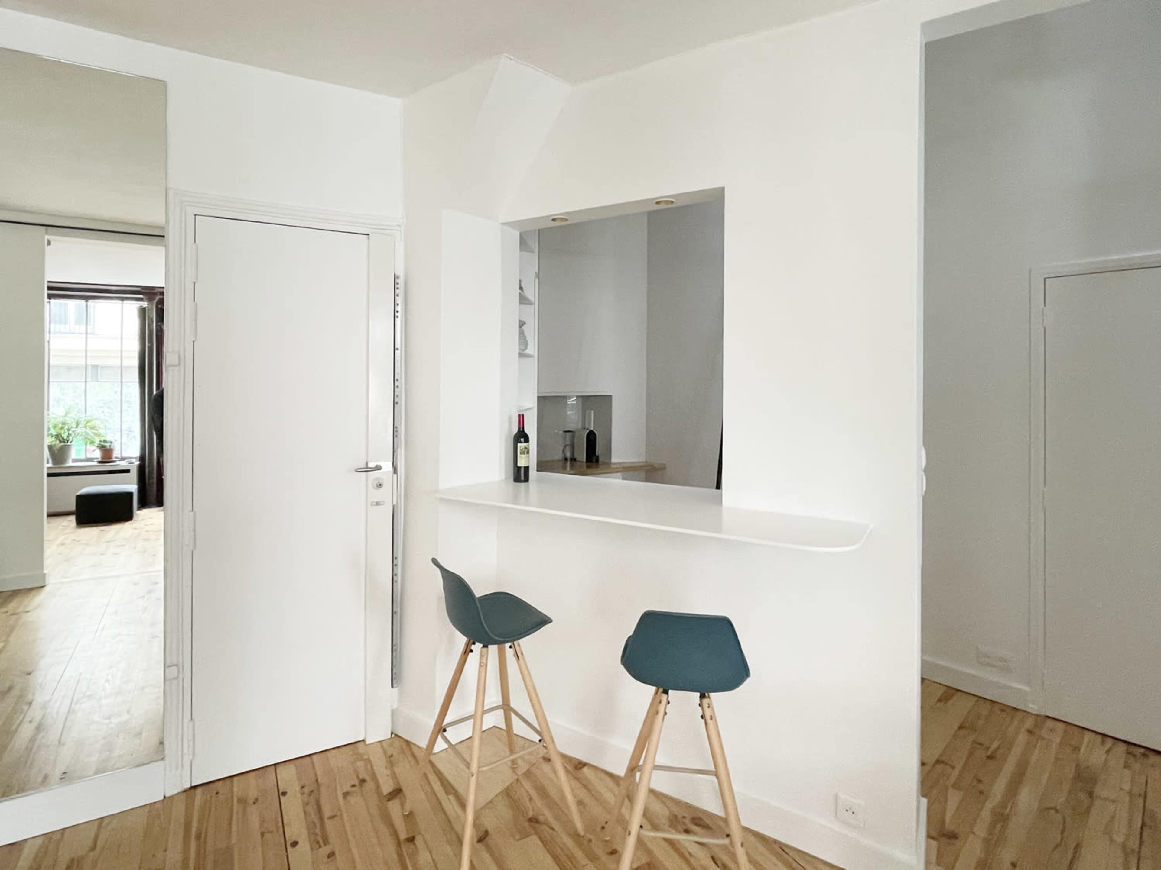 A minimalist interior space featuring a small bar area with two stools, a white wall, and a door leading to another room.