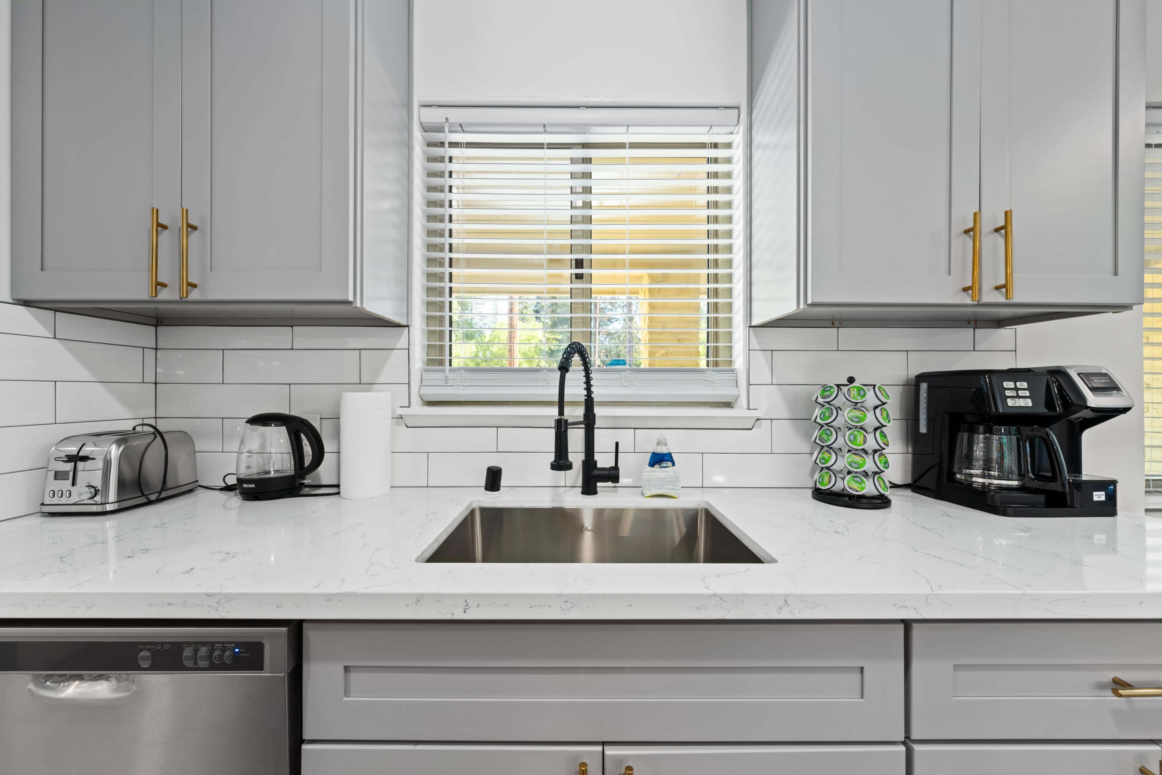 A modern kitchen countertop with a sink, coffee maker, toaster, and various kitchen items neatly arranged against light gray cabinets.