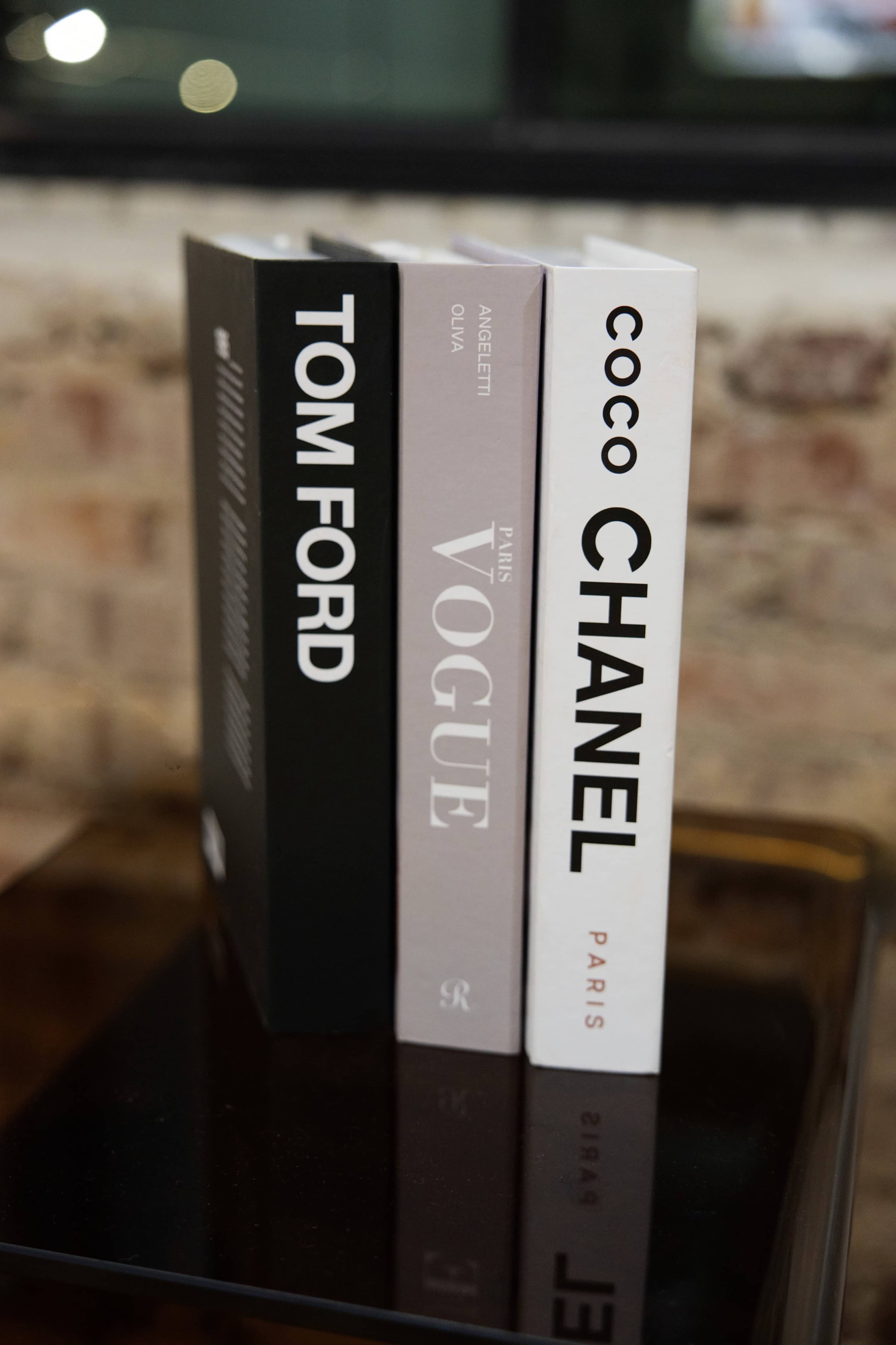The image shows three stylish hardcover books titled "TOM FORD," "VOGUE," and "COCO CHANEL," arranged on a reflective surface with a brick wall in the background.