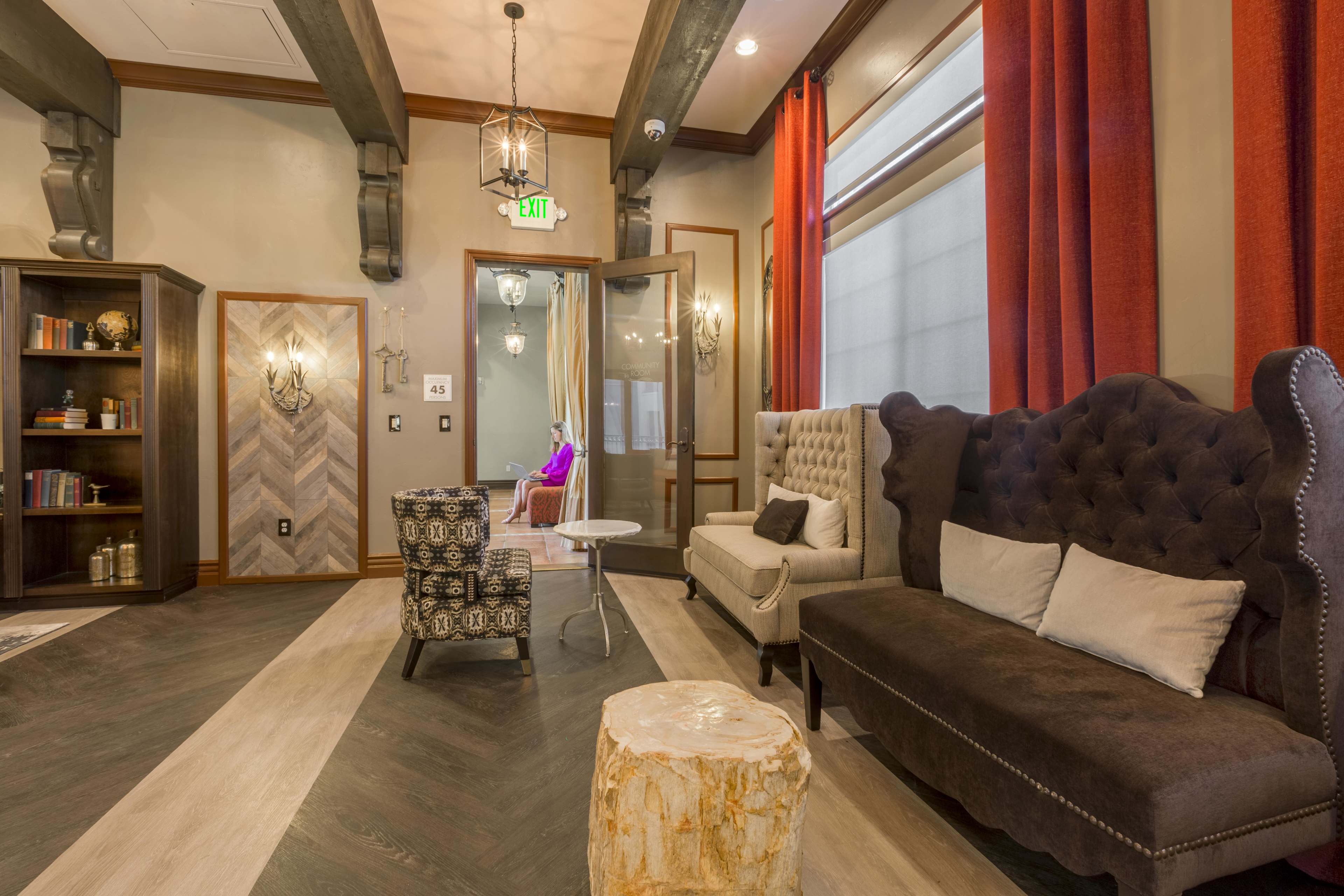 The image shows a stylish interior with two upholstered sofas, a decorative armchair, a small white table, and a wooden stump, all placed on a patterned floor with large windows and red drapes.