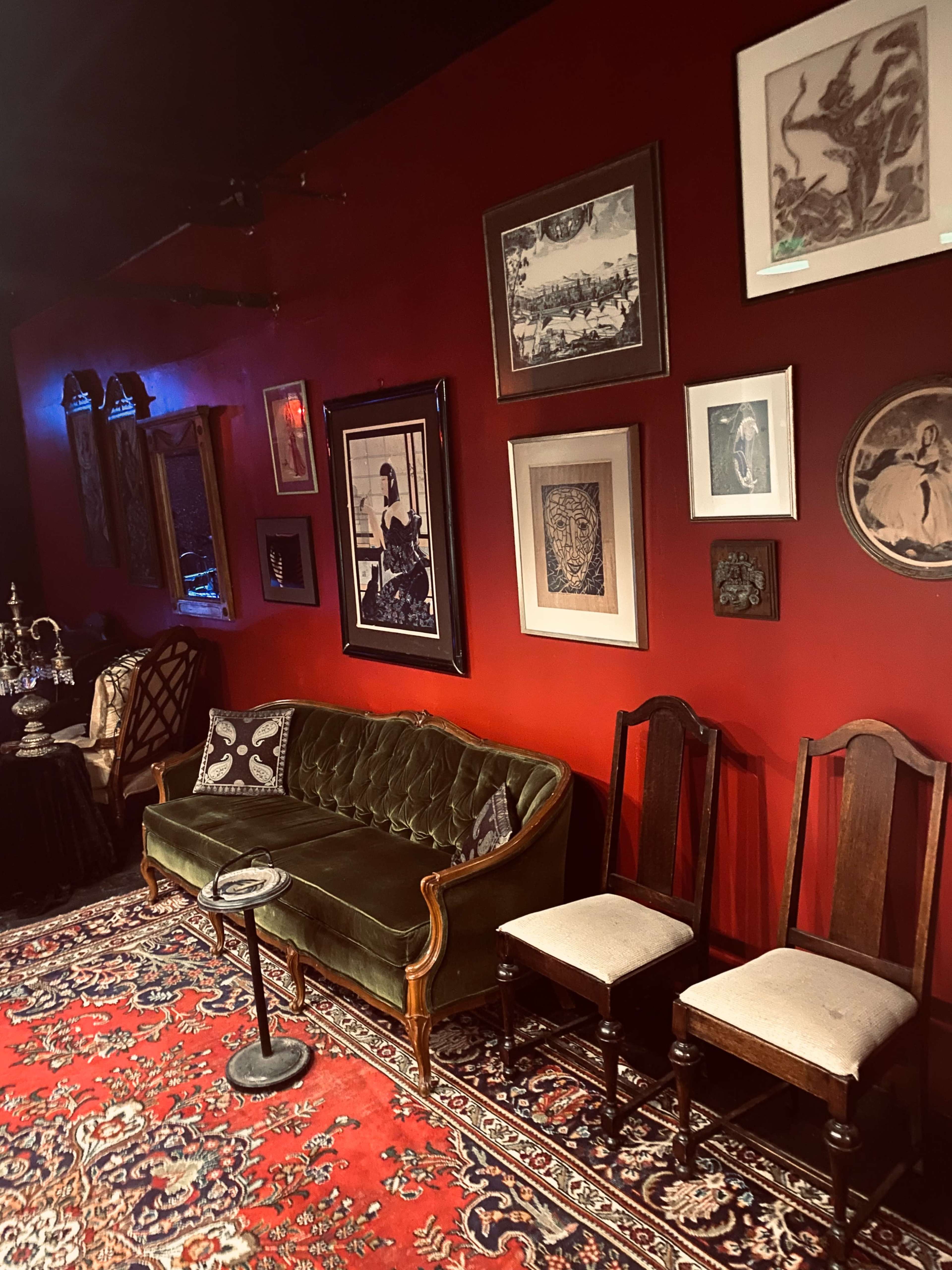 The image shows a cozy, dimly lit room with a green velvet sofa and two wooden chairs against a deep red wall adorned with various framed artworks.