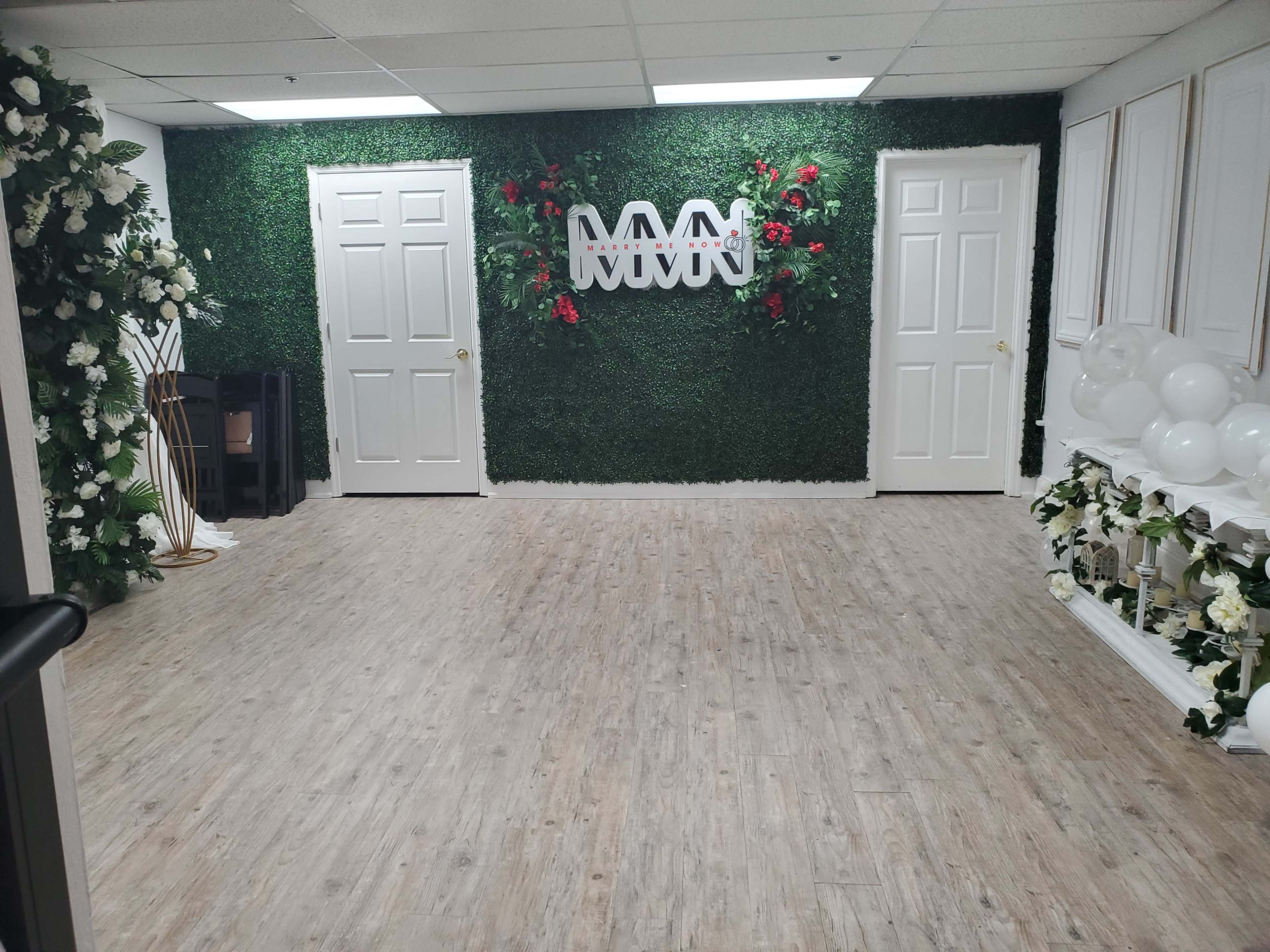 The image shows a well-decorated room featuring green hedge walls, white doors, floral arrangements, and a white balloon display.