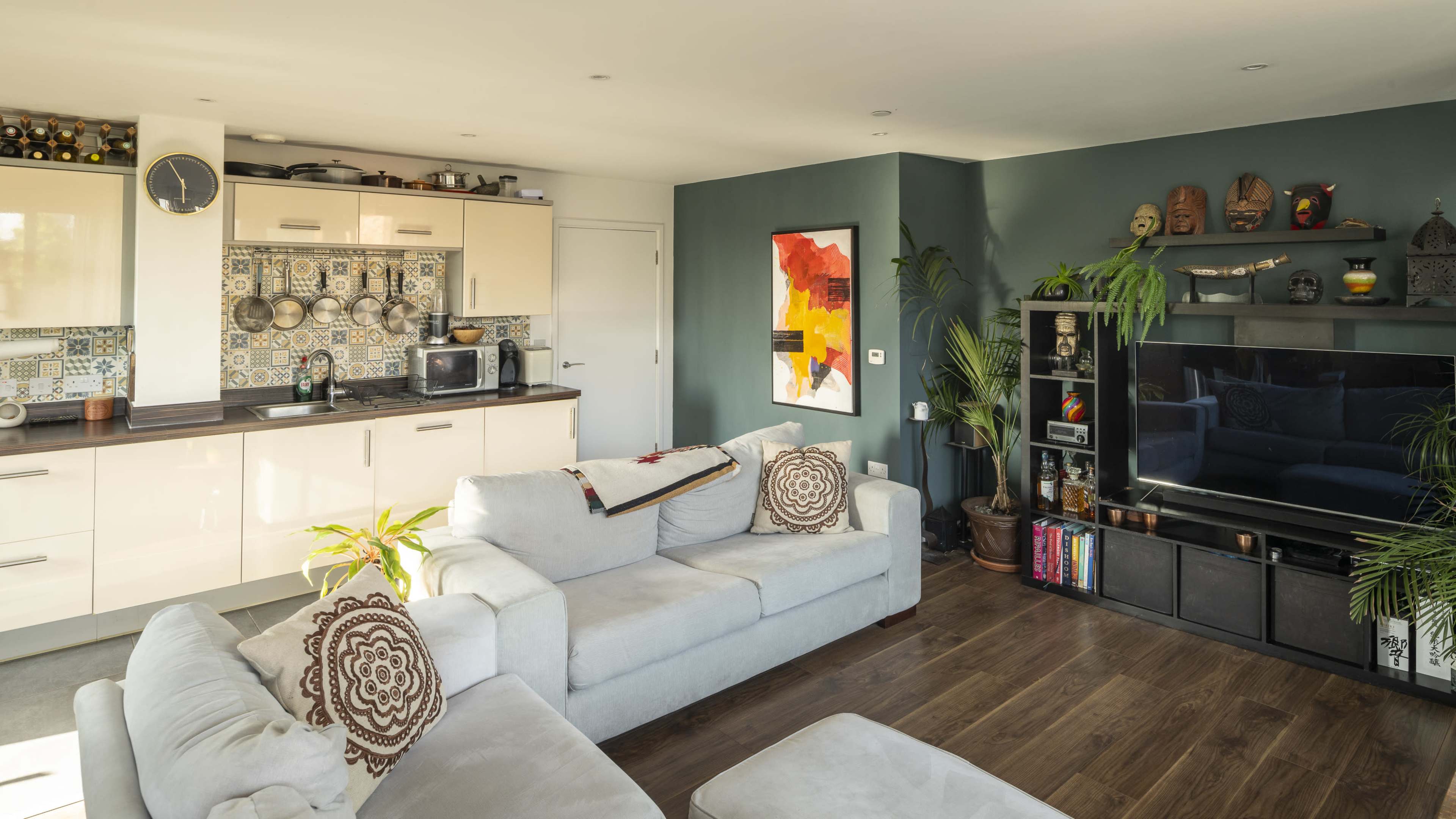 The image shows a modern living room with a light-colored sectional sofa, a large television, and a kitchen area featuring beige cabinets and colorful decorative tiles.