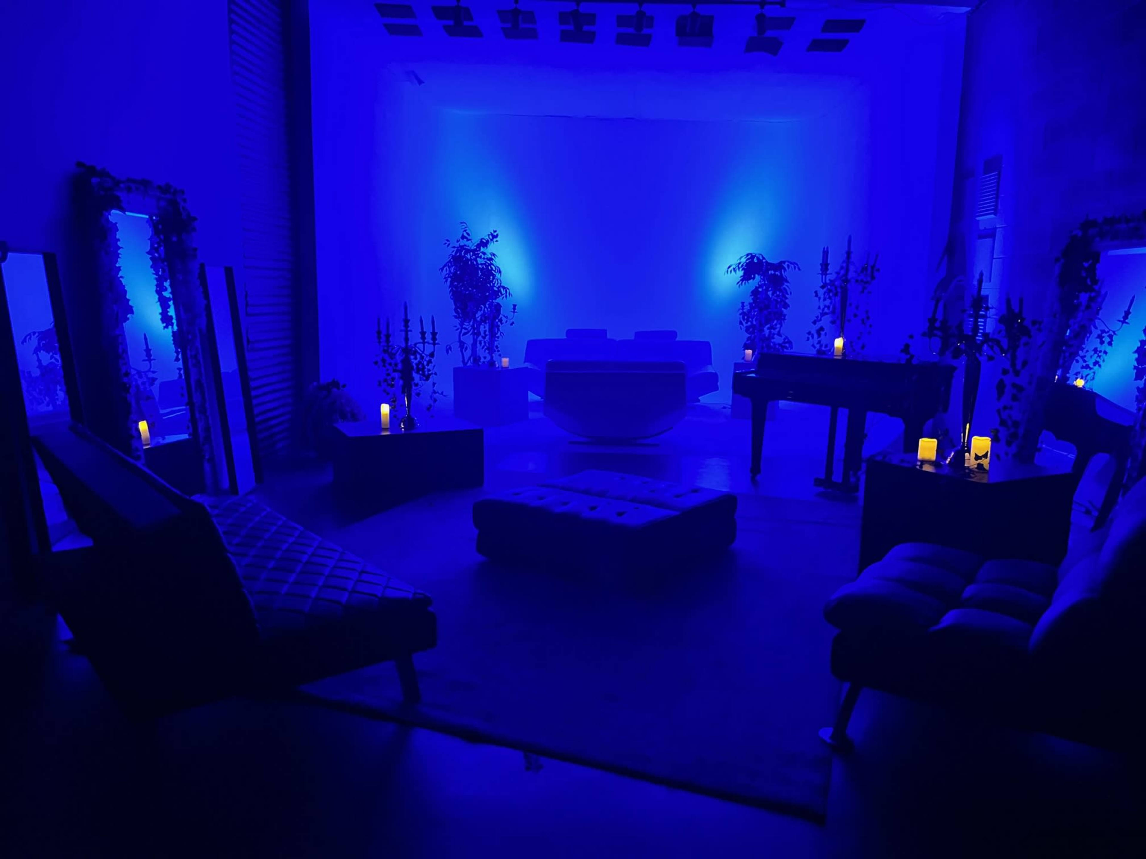 The image shows a dimly lit room with blue lighting, featuring various furniture pieces, plants, and candles arranged throughout the space.
