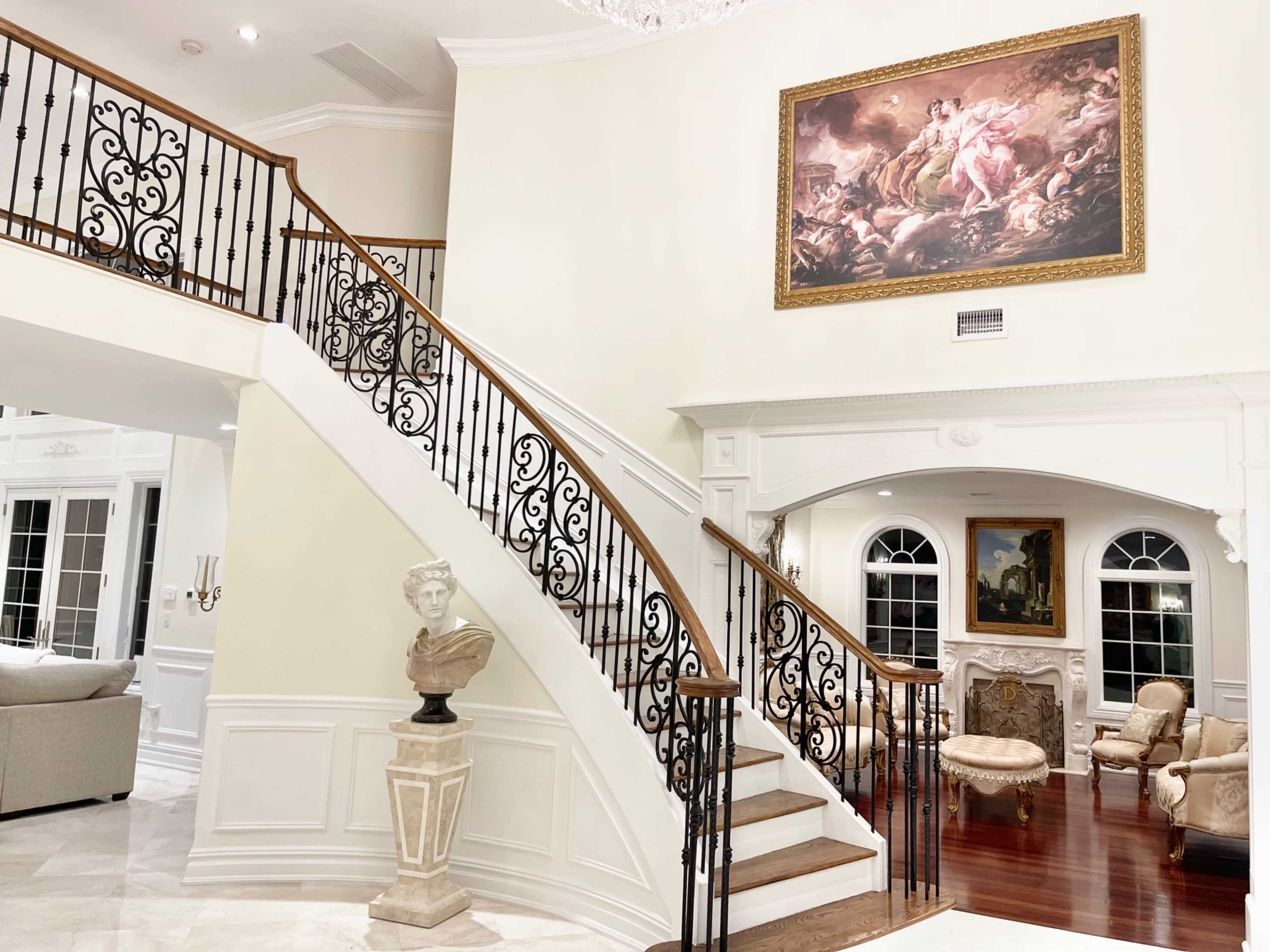 An elegant foyer featuring a grand staircase with ornate black railings, a large painting on the wall, and classic furniture in a well-lit space.