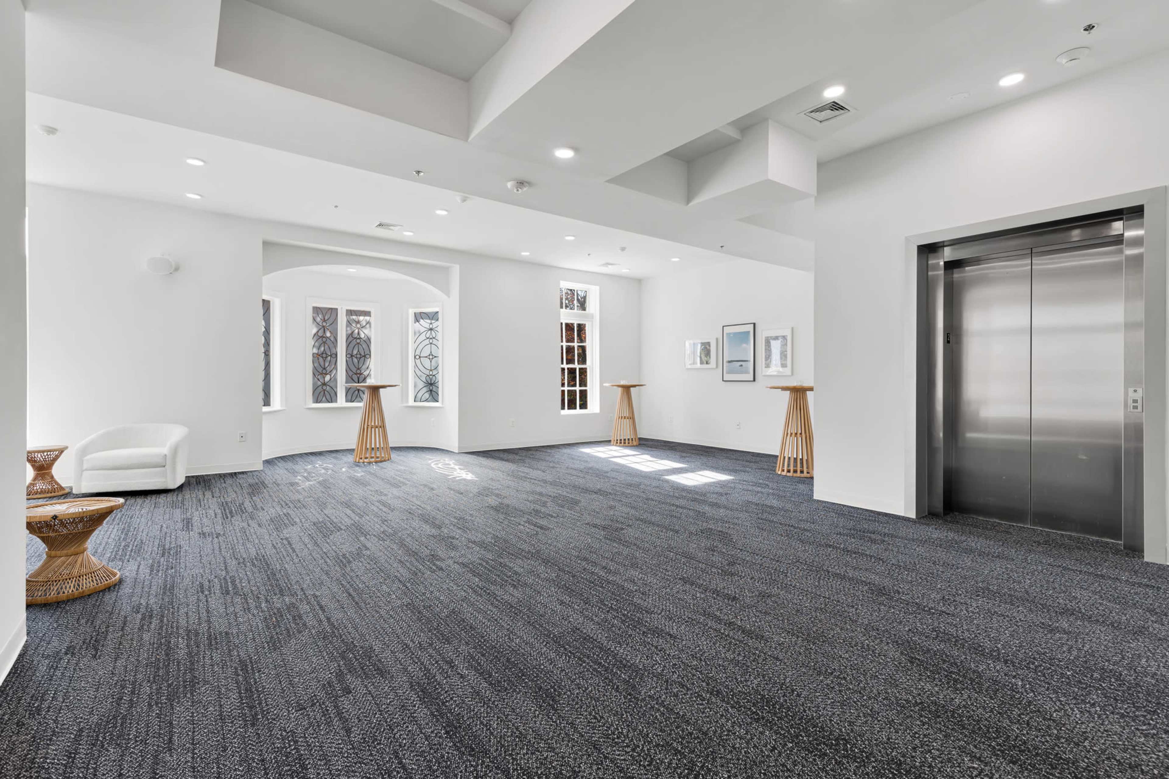 The image shows a spacious, well-lit interior with a carpeted floor, an elevator, and minimal furniture, including stools and a chair.