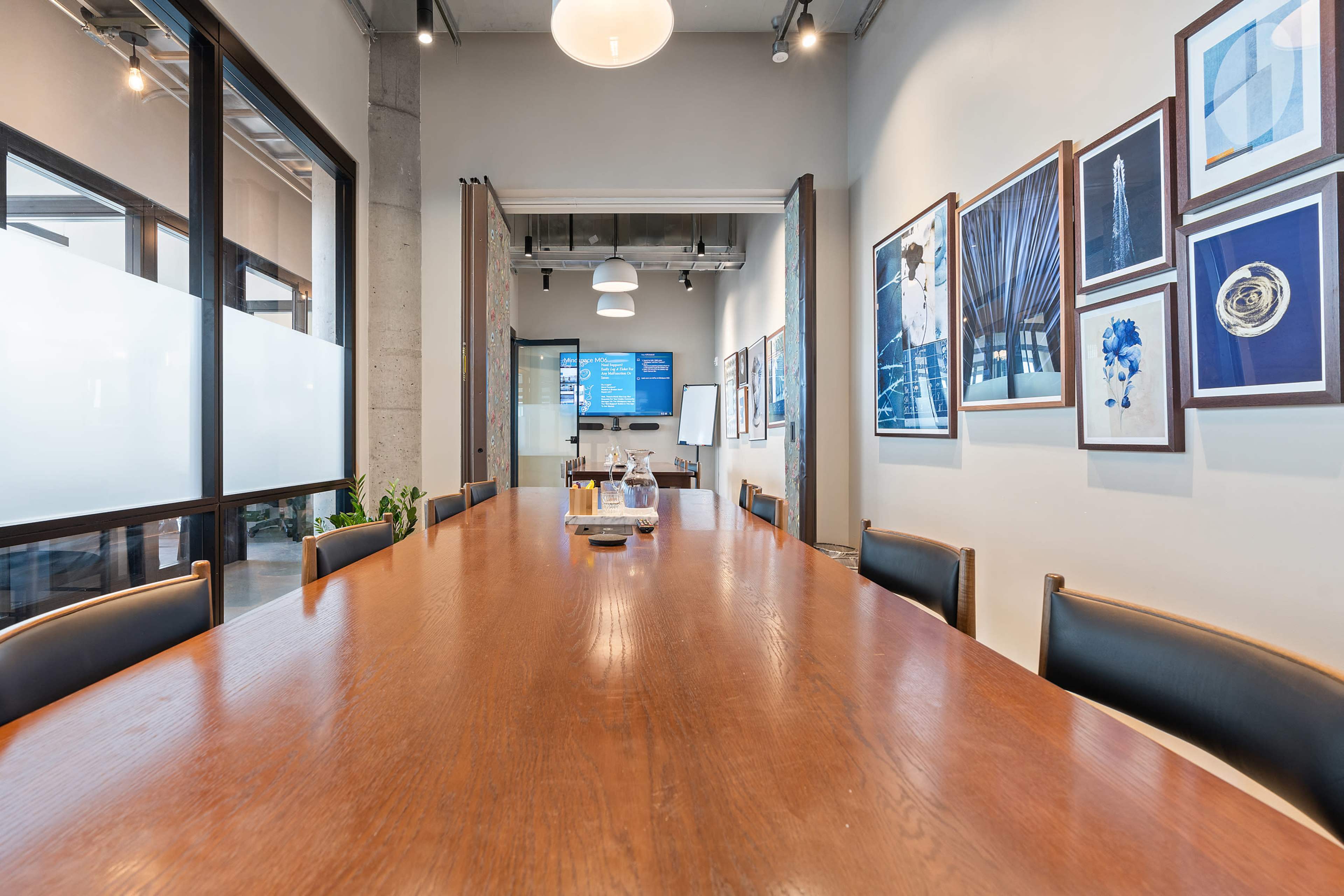 A long wooden conference table is surrounded by chairs in a modern meeting room, featuring artwork on the walls and a screen at the far end.