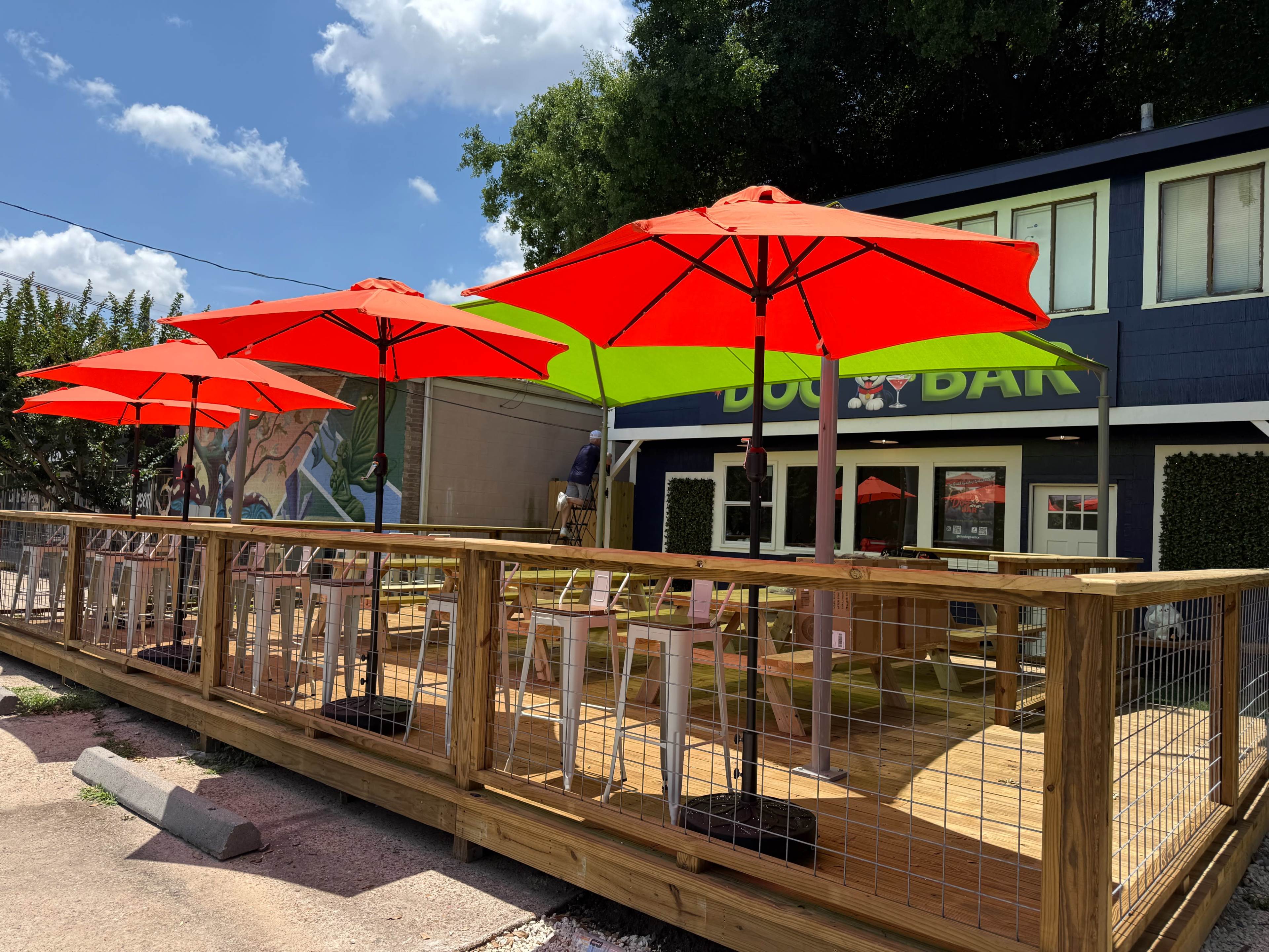 The image shows a wooden deck of a bar with bright red and green umbrellas providing shade over outdoor seating.