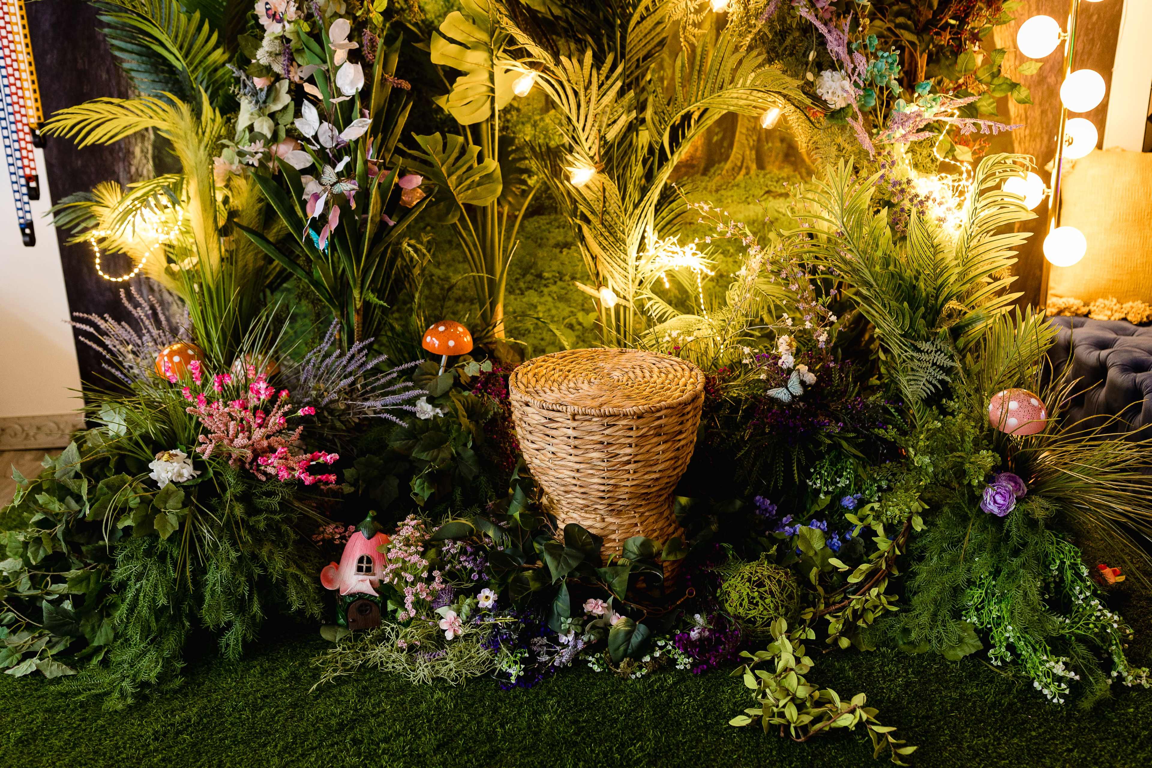 A decorated area with lush greenery, colorful flowers, and a woven basket surrounded by various plants and ambient lighting.