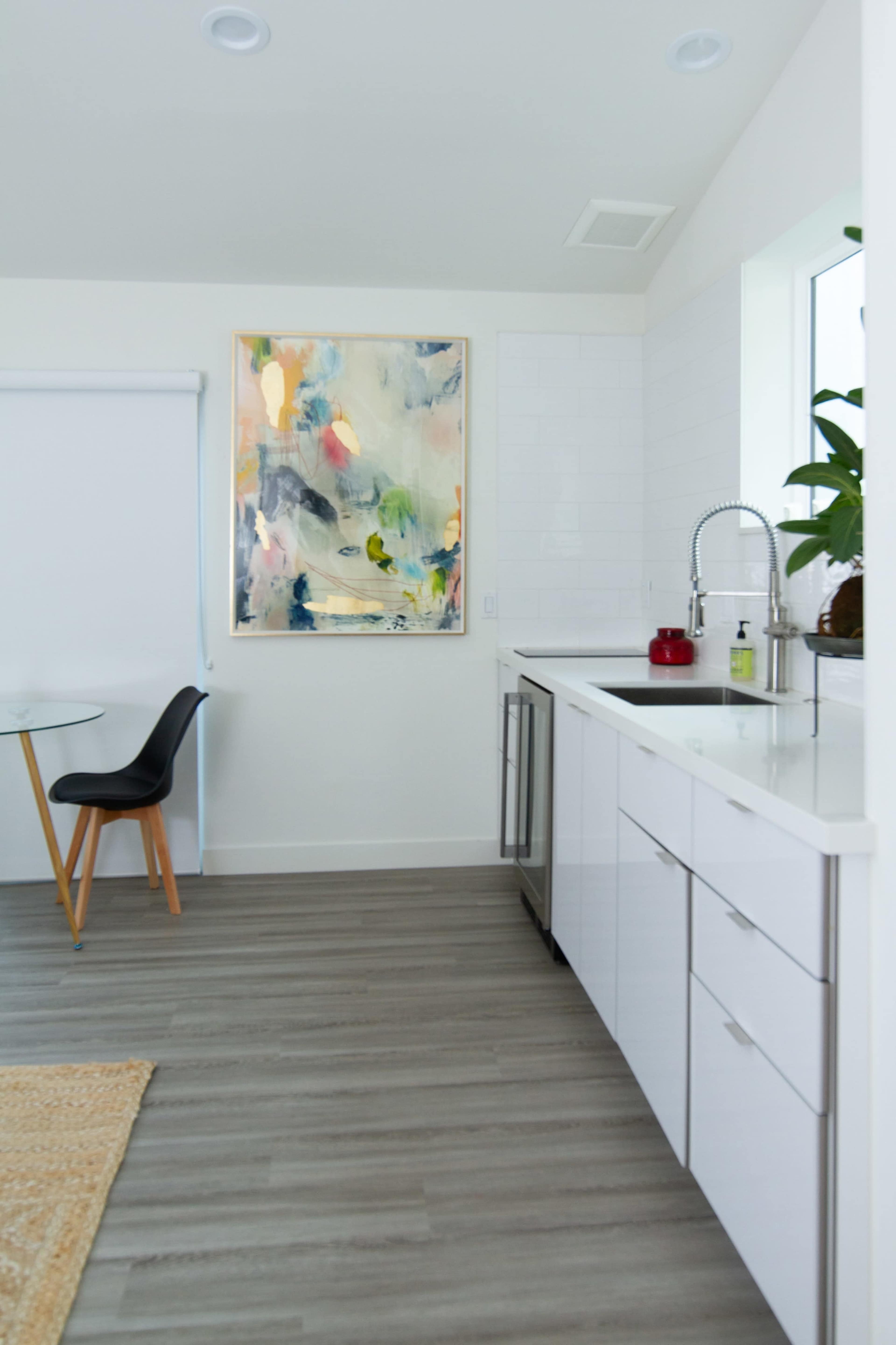 A modern kitchen features a stainless steel sink, white cabinetry, a round dining table with a black chair, and a colorful abstract painting on the wall.