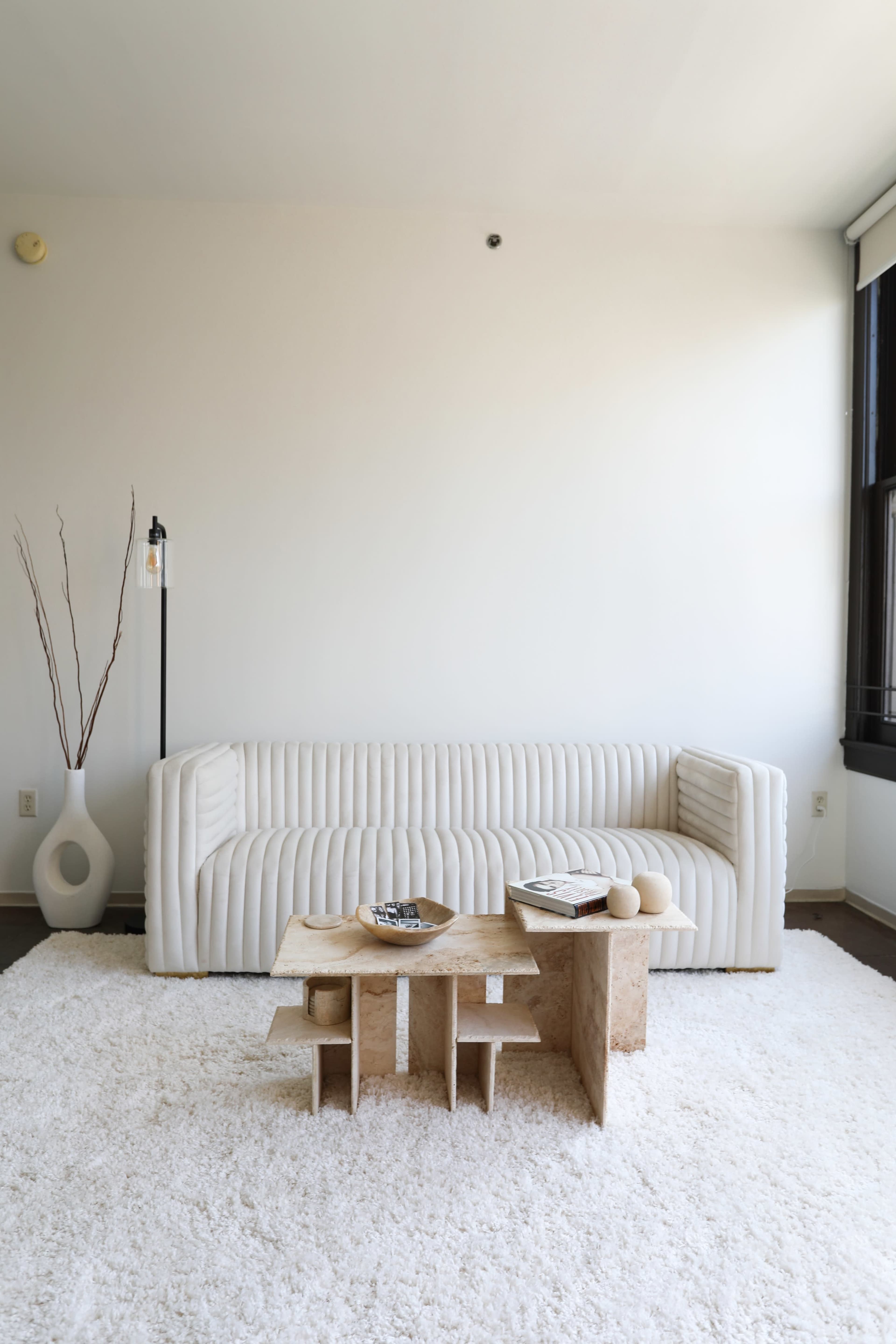 A modern, minimalist living room features a white ribbed sofa, a light-colored textured coffee table, and a soft area rug.
