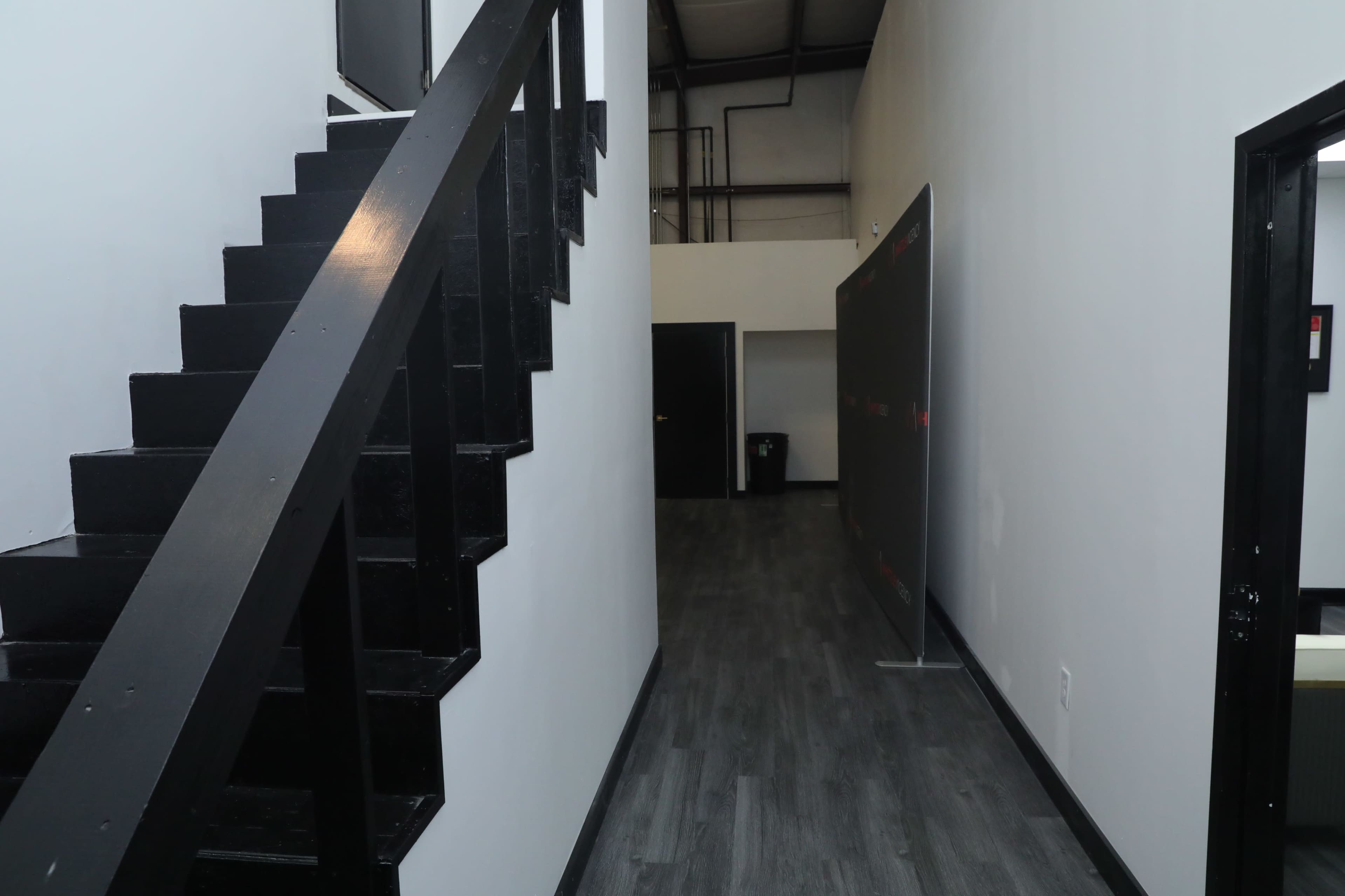 A narrow hallway with black stairs on the left and a partition wall on the right, leading to a door at the end.