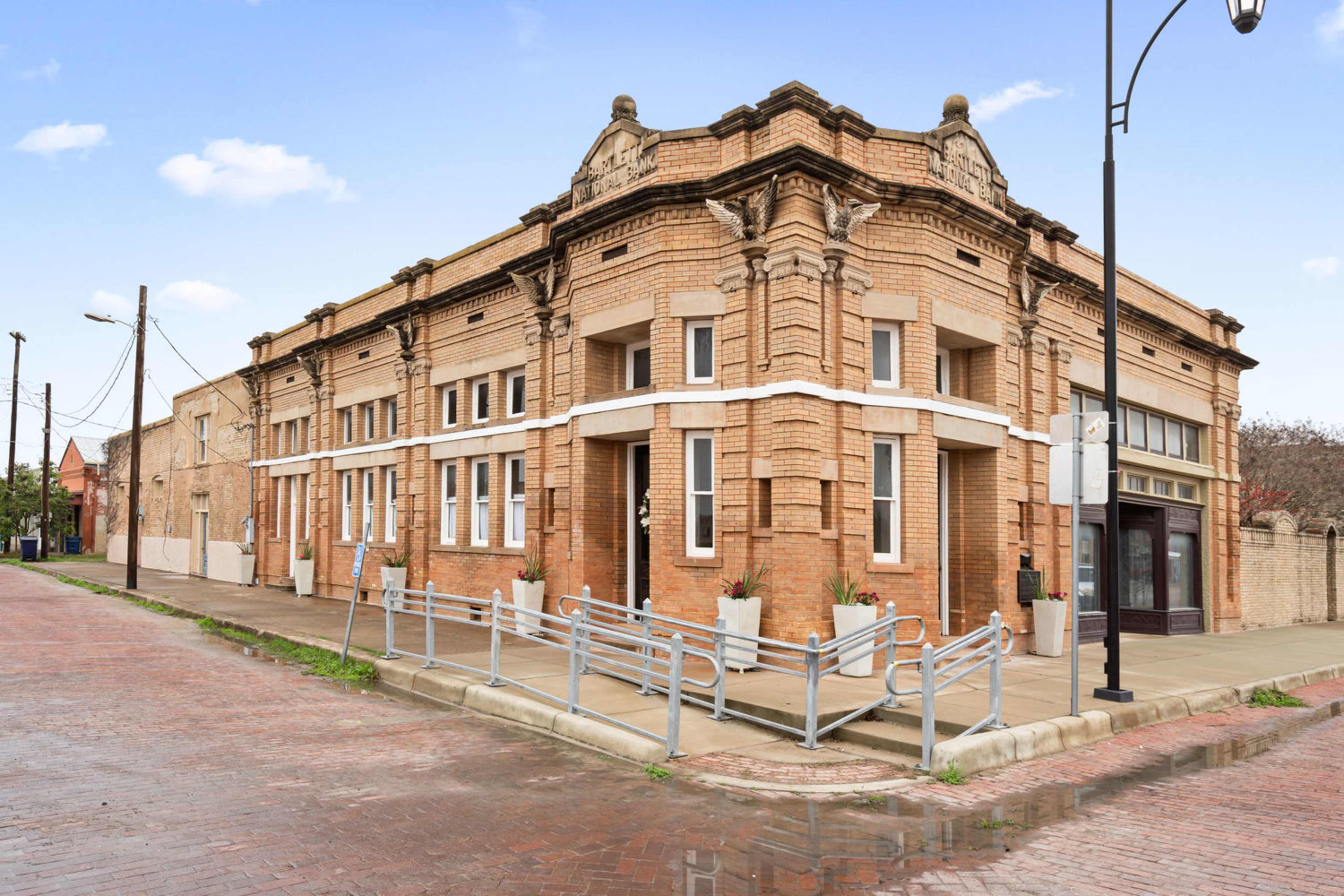 A brick building with architectural details at the intersection of two streets, surrounded by a paved area and street lamps.