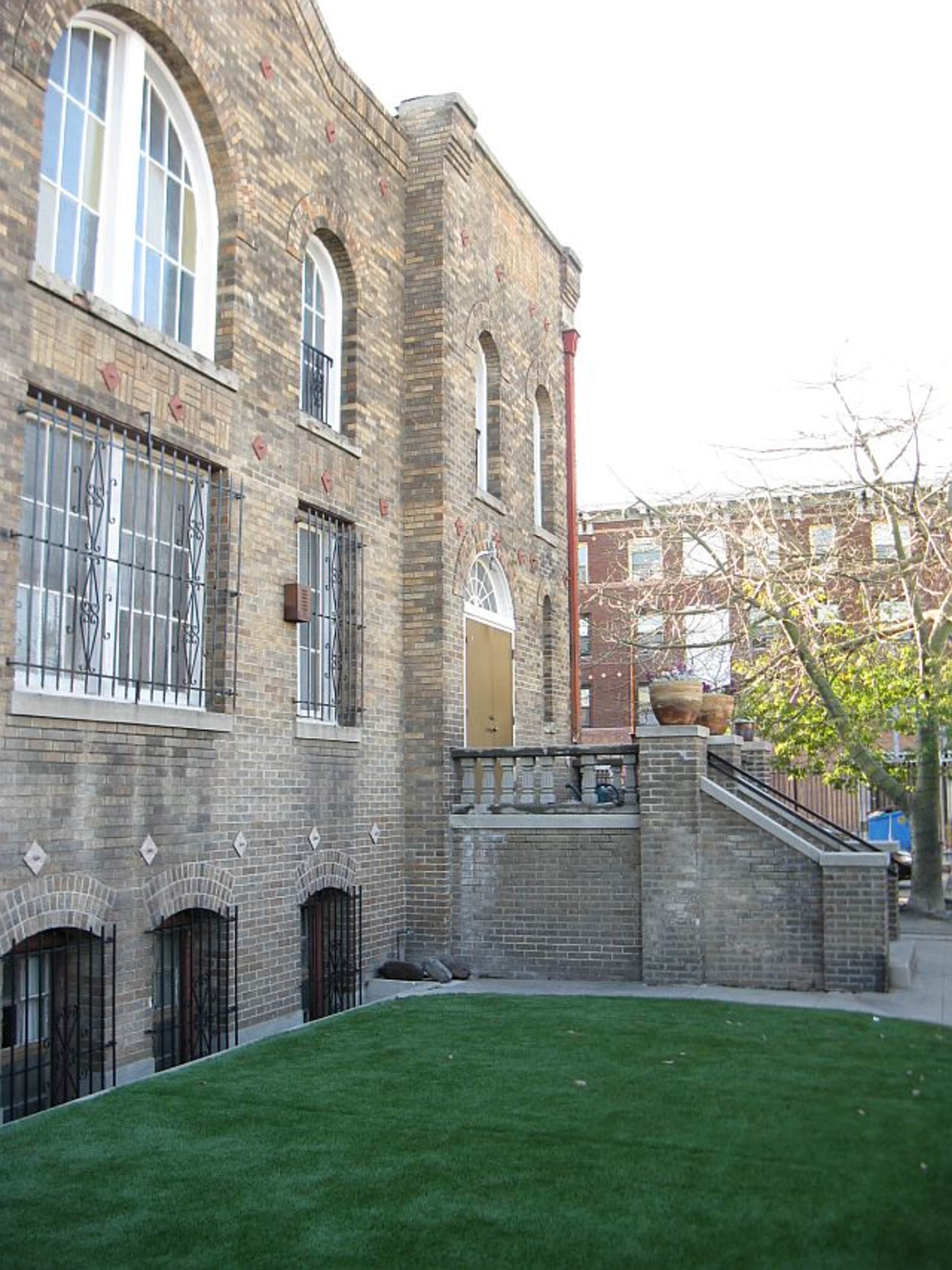 A brick building with arched windows and a staircase leading to a large door, surrounded by a small lawn area.