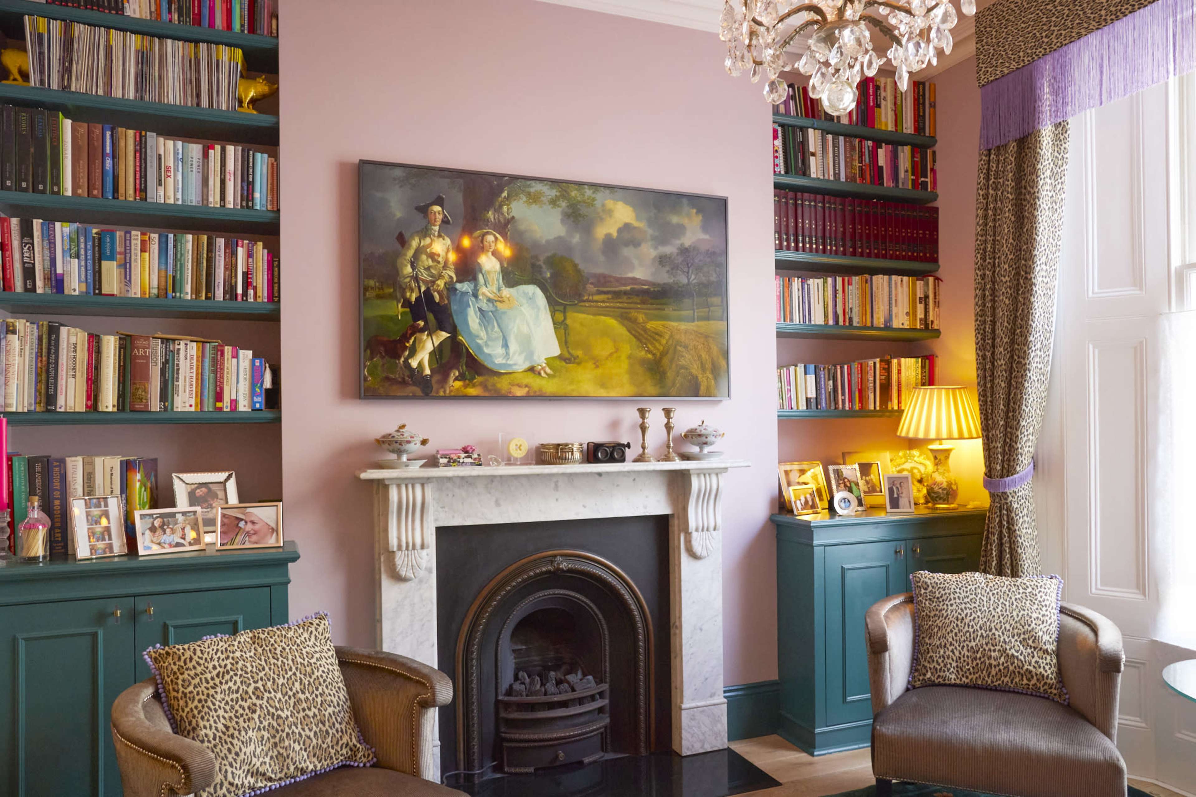 A cozy living room with bookshelves on either side, a marble fireplace beneath a painting, and two armchairs arranged in front of a coffee table.