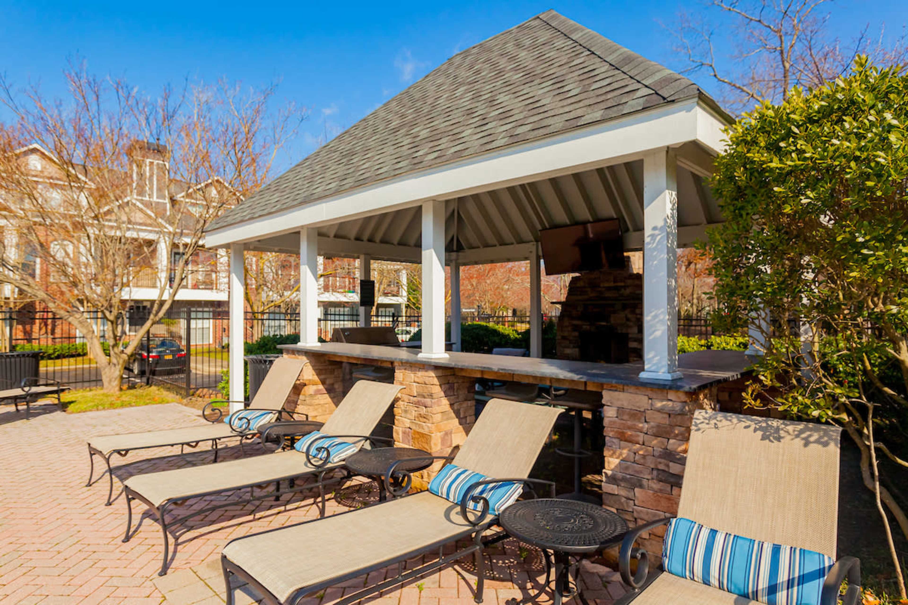 A shaded outdoor pavilion with a stone bar area next to several lounge chairs arranged around a paved patio.