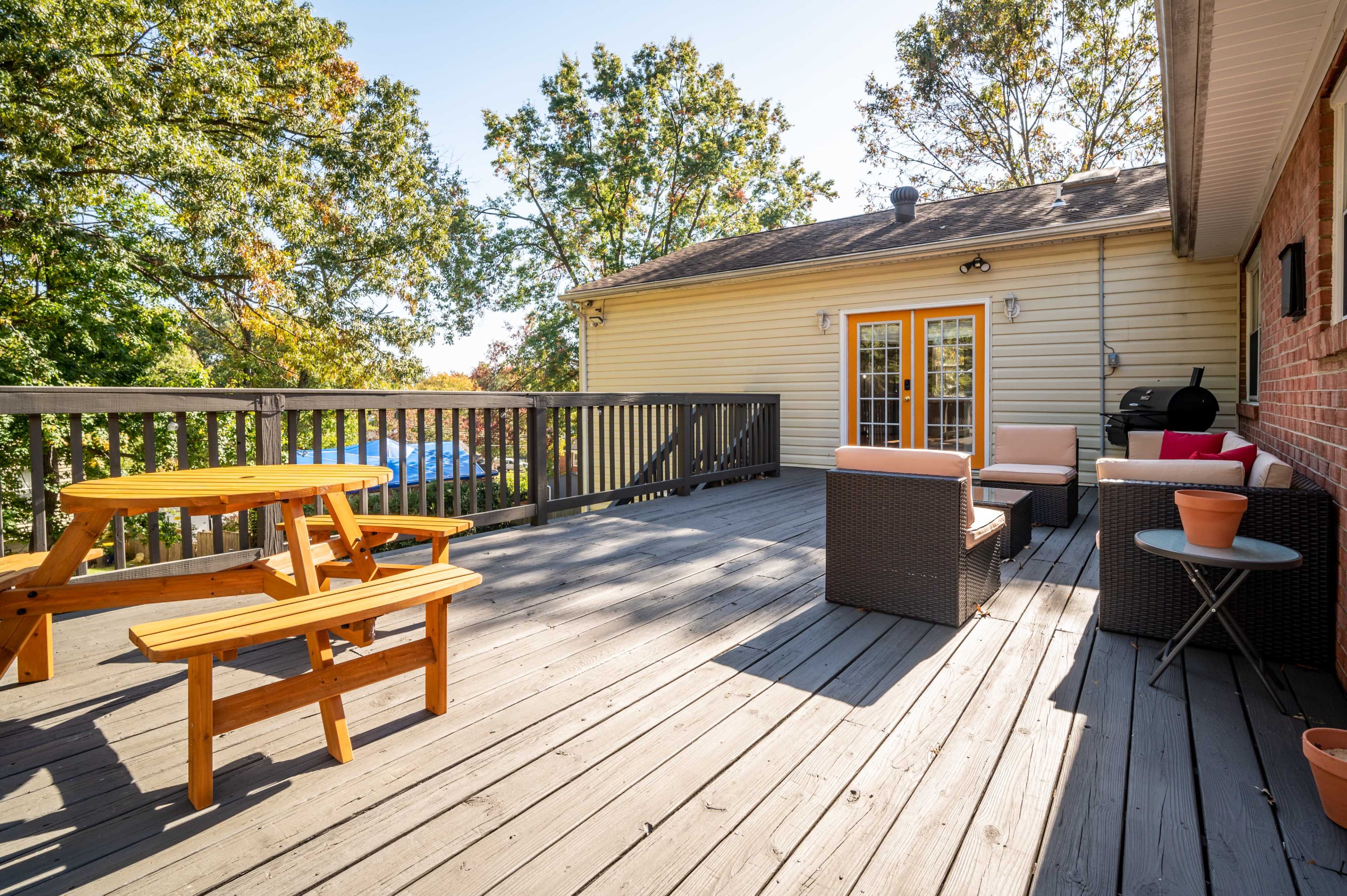 A wooden deck features a picnic table and seating area with cushions, surrounded by trees and a grill nearby.