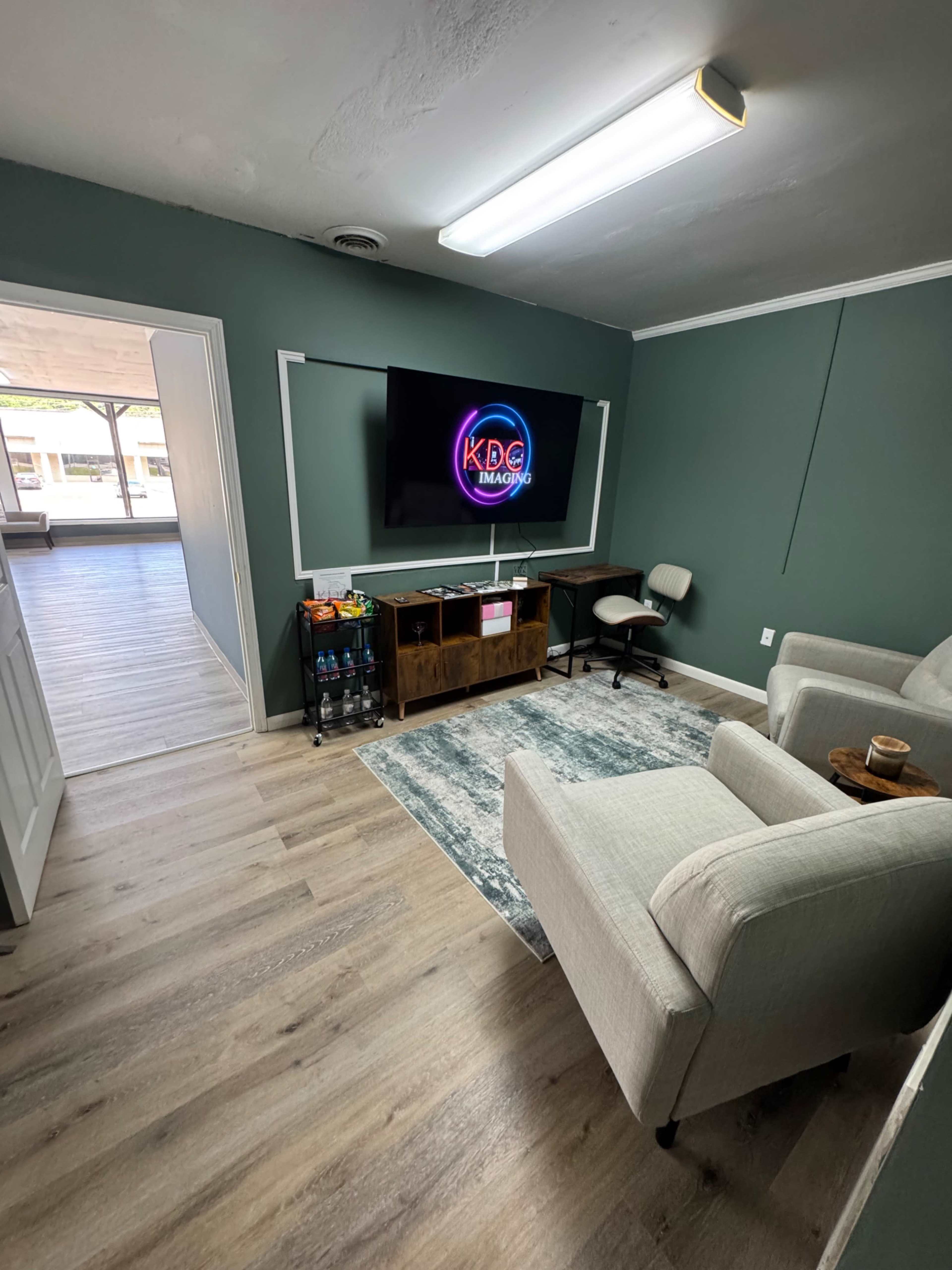 The image shows a cozy waiting area with a television mounted on the wall, a grey armchair, a small table with snacks, and a rug on a wooden floor.