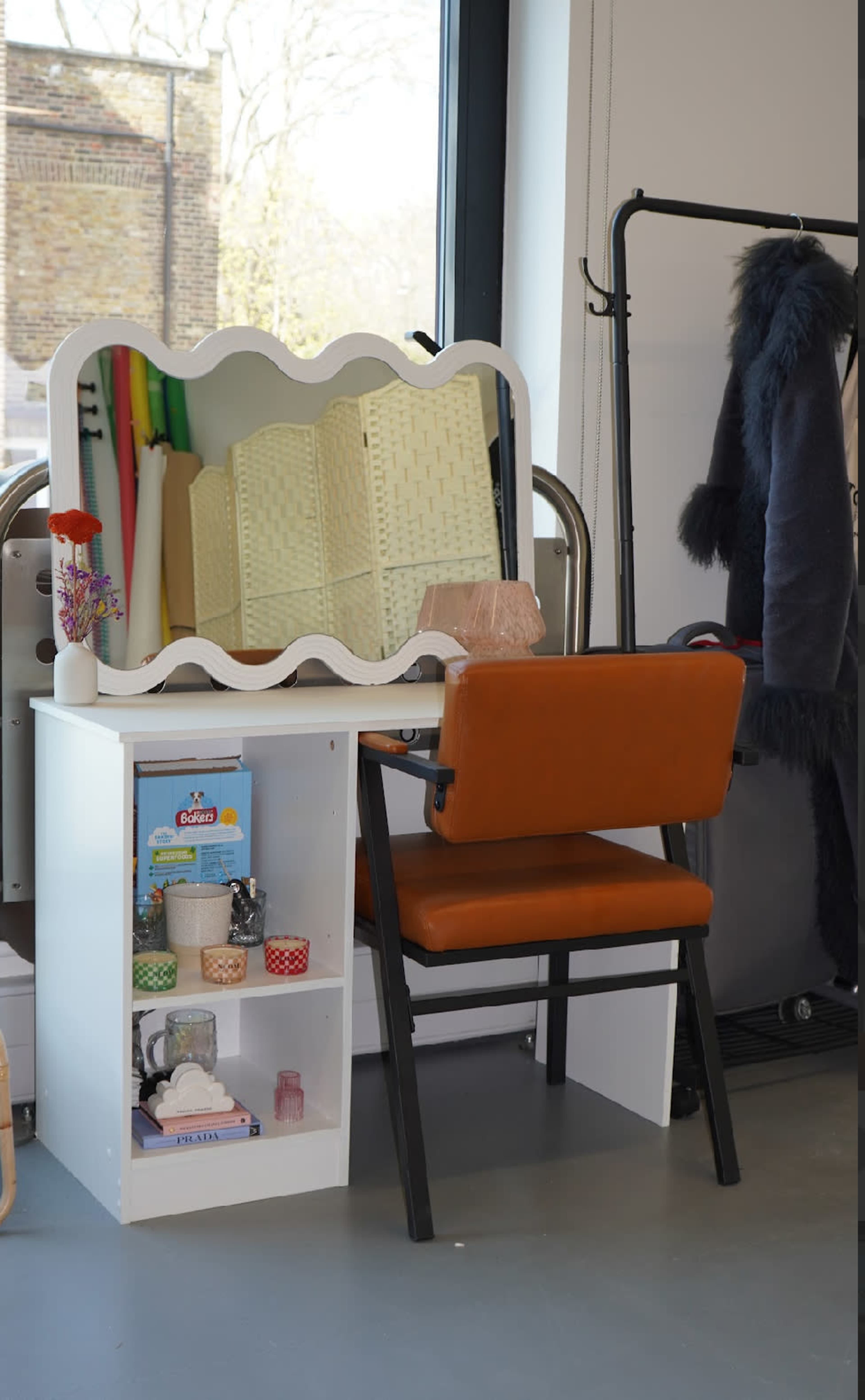 The image shows a white vanity table with a scalloped mirror, accompanied by an orange chair and various items displayed on the table and shelf.