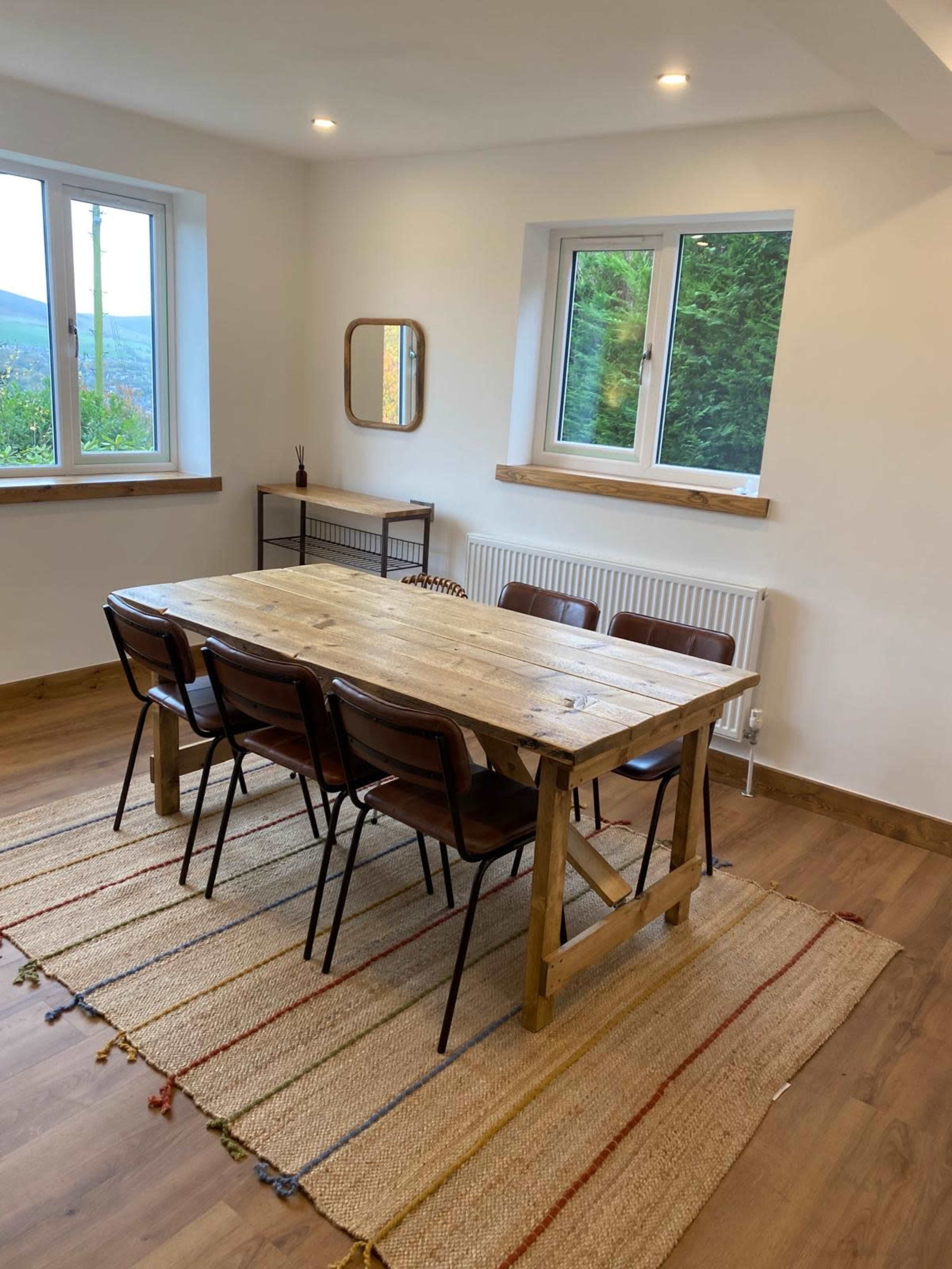 A rustic wooden dining table is surrounded by six chairs on a patterned rug, situated in a well-lit room with two windows.
