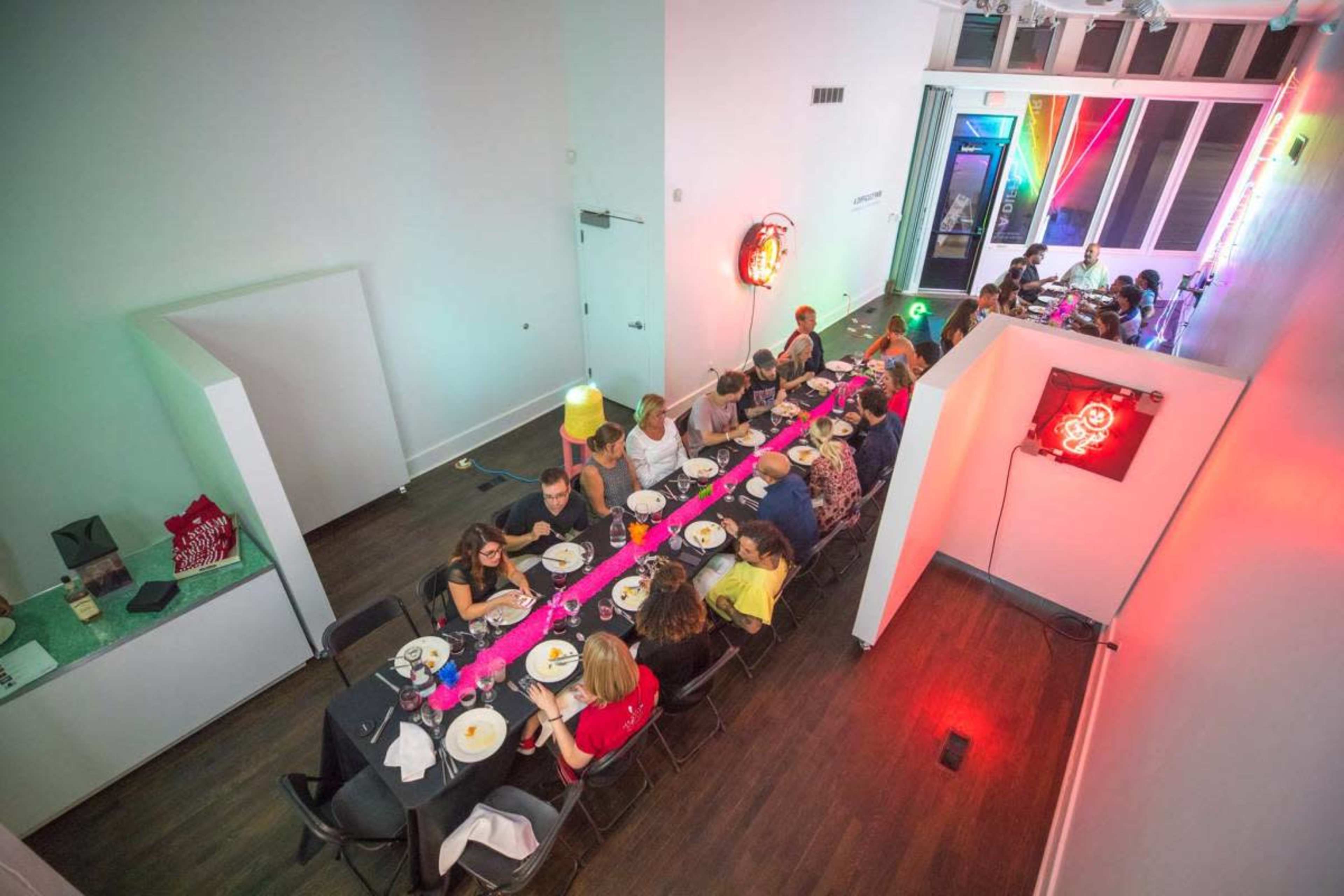 A long dining table is set with plates and drinks, while guests are seated along it in a brightly lit modern space.