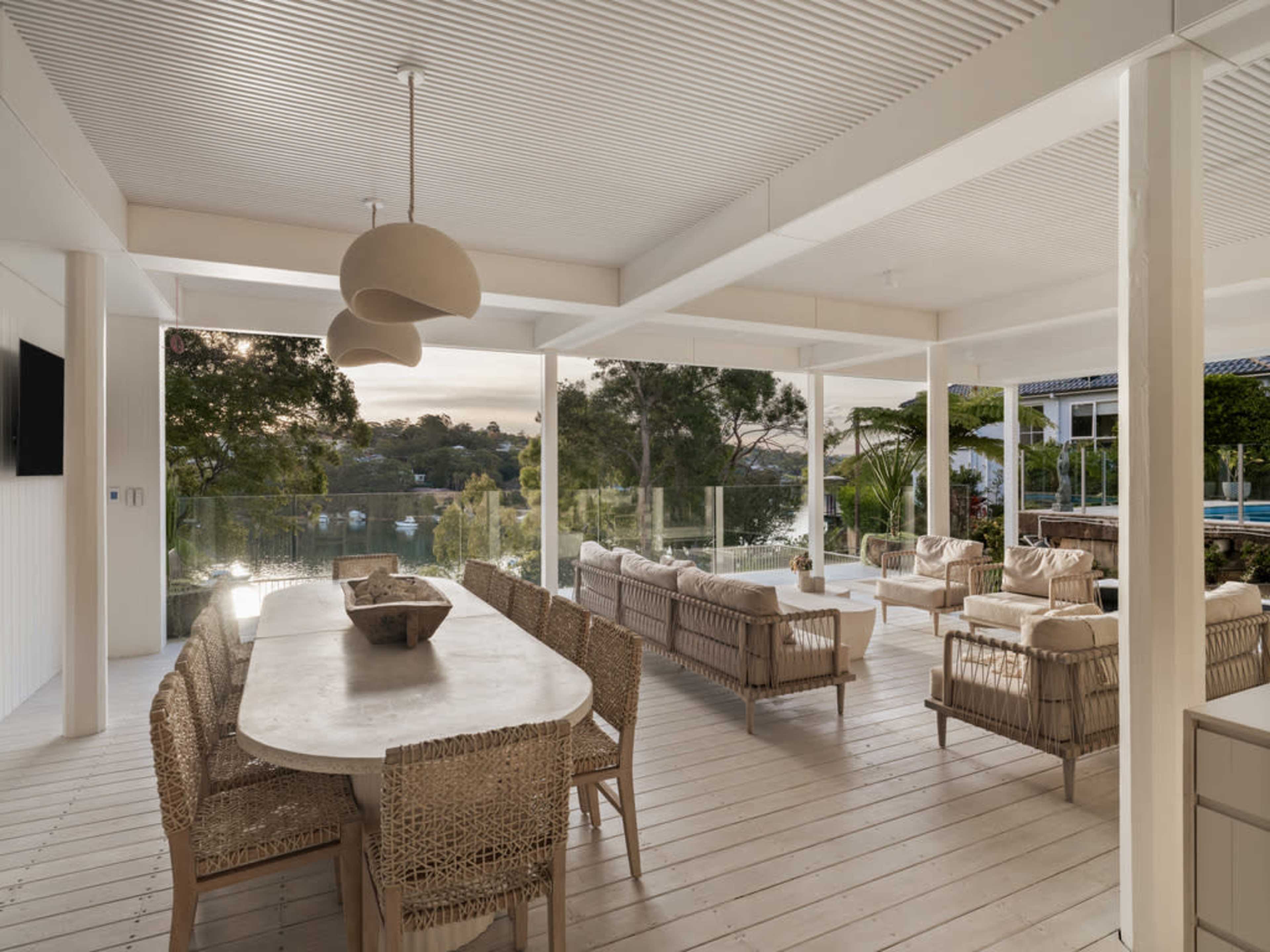 The image shows a modern outdoor patio area featuring a large dining table with wicker chairs and a seating arrangement, surrounded by greenery and overlooking a body of water.