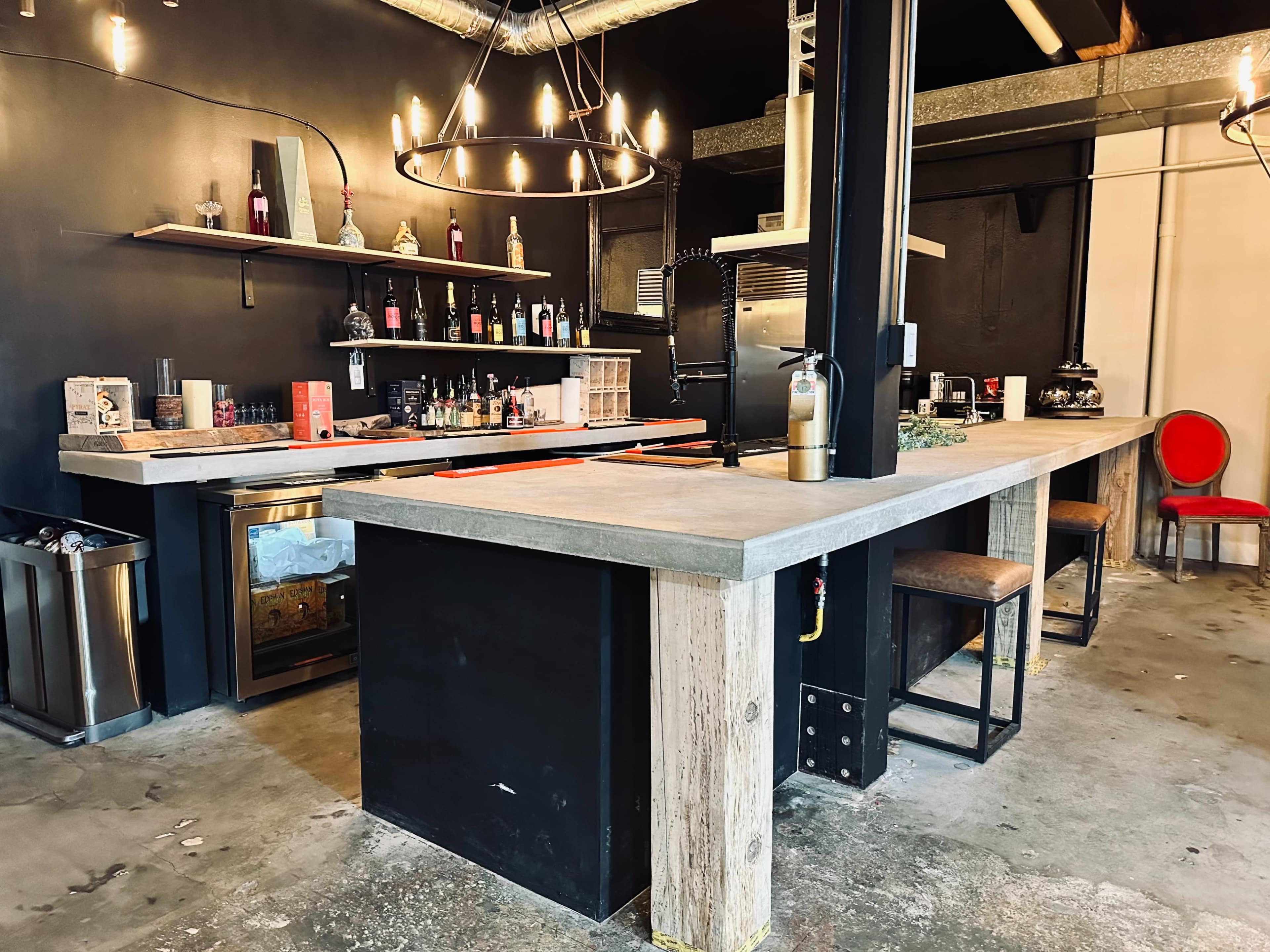 The image shows a modern bar area with a concrete countertop, black walls, a shelf stocked with bottles, and wooden stools.