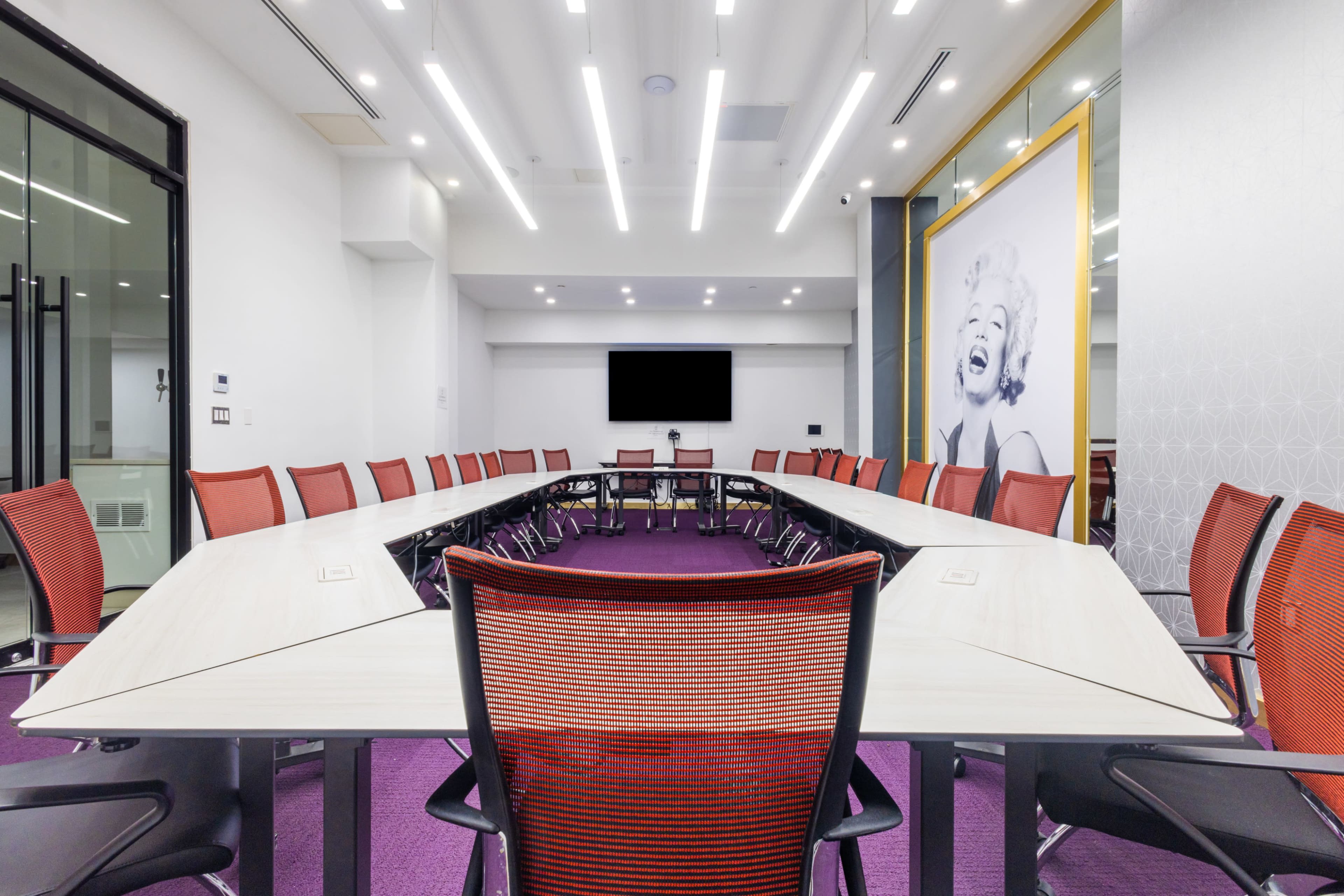 A conference room features a long table surrounded by orange chairs, with a large screen at one end and a wall displaying a large image of a woman.