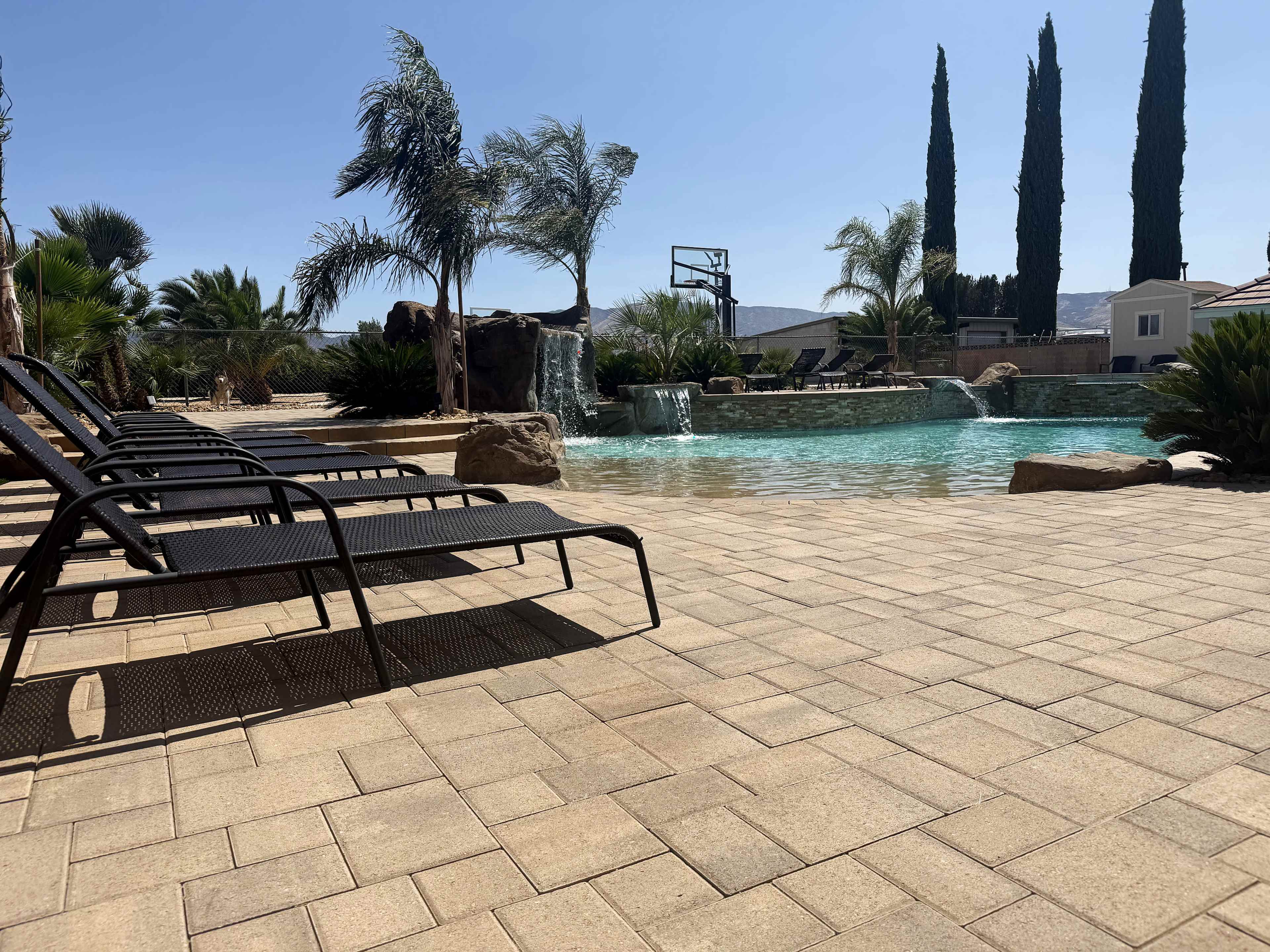 The image shows a swimming pool area with lounge chairs, palm trees, a waterfall feature, and a basketball hoop in the background.