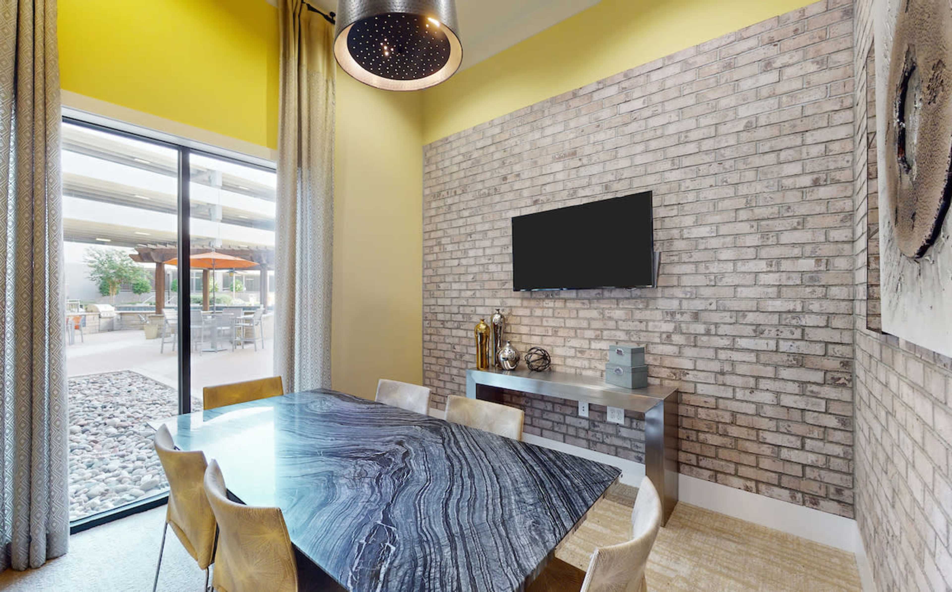 A modern dining area features a sleek table under a pendant light, with a large window showcasing an outdoor view and a TV mounted on the brick wall.