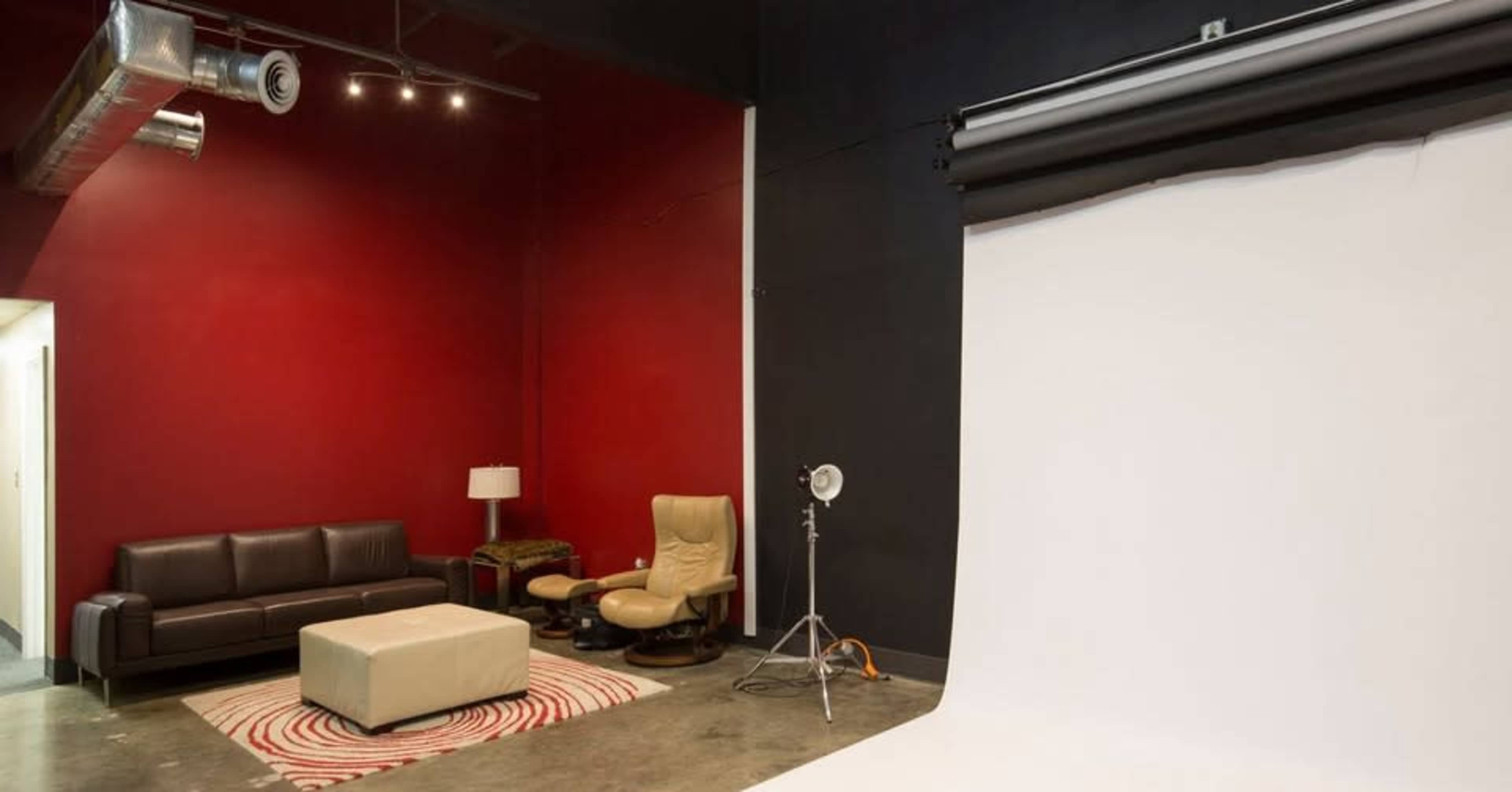 The image shows a studio space with a red wall on one side, a white backdrop set up for photography, a brown couch, an ottoman, and a floor lamp.