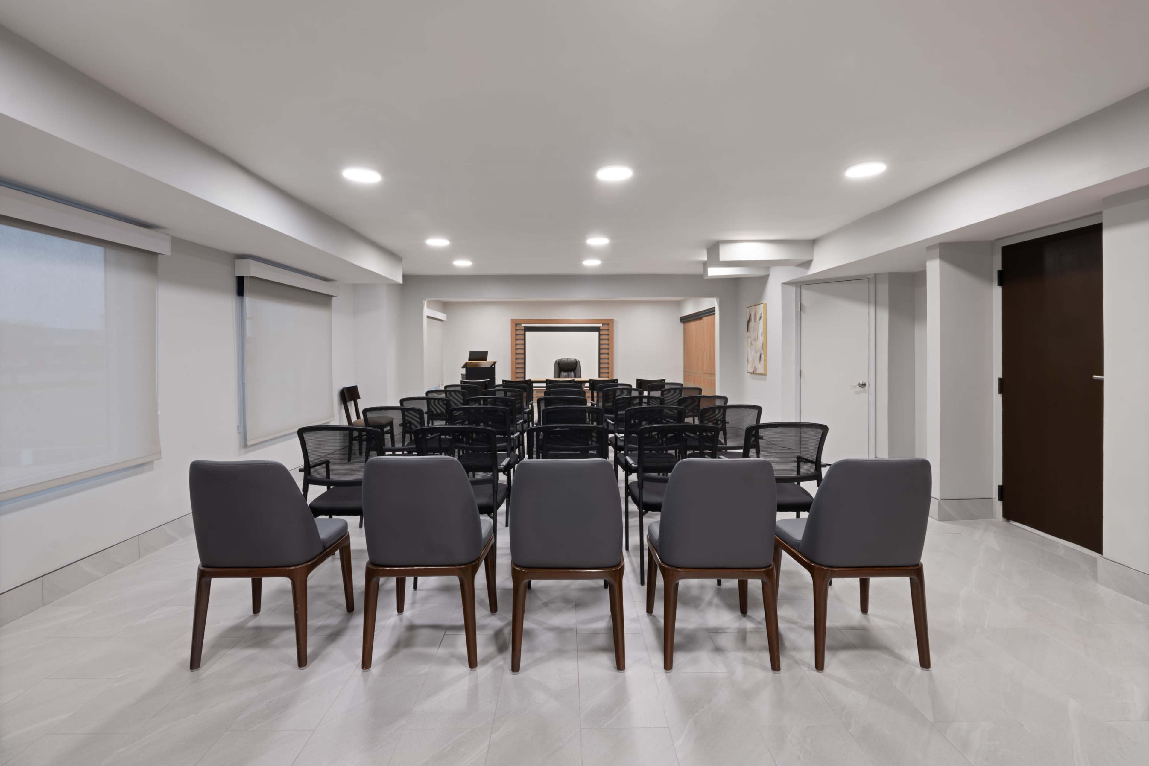 The image shows a conference room with rows of black and gray chairs facing a podium and a large wall-mounted mirror.