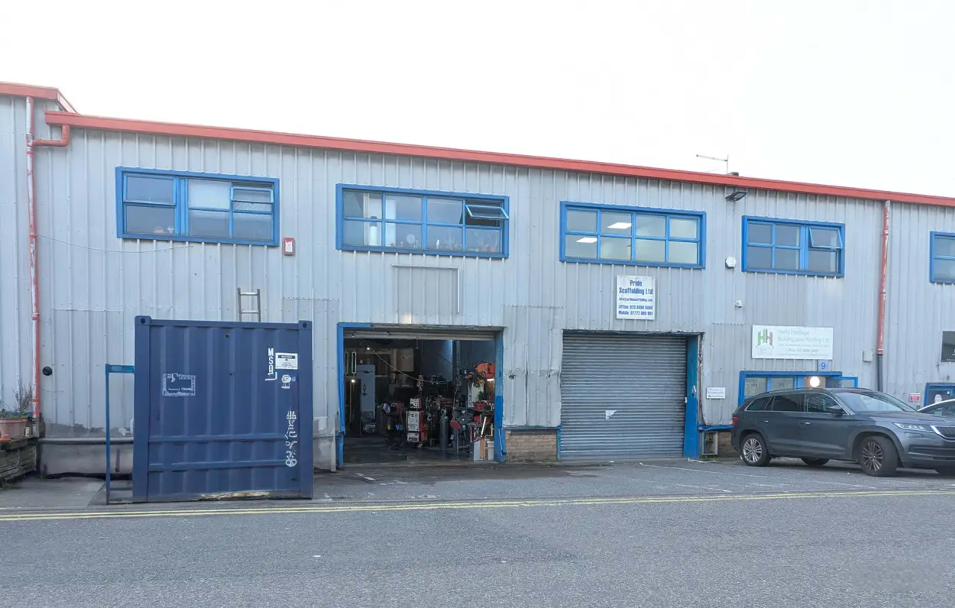 The image shows a gray metal building with blue windows and a loading dock, featuring an open entrance and parked vehicles outside.