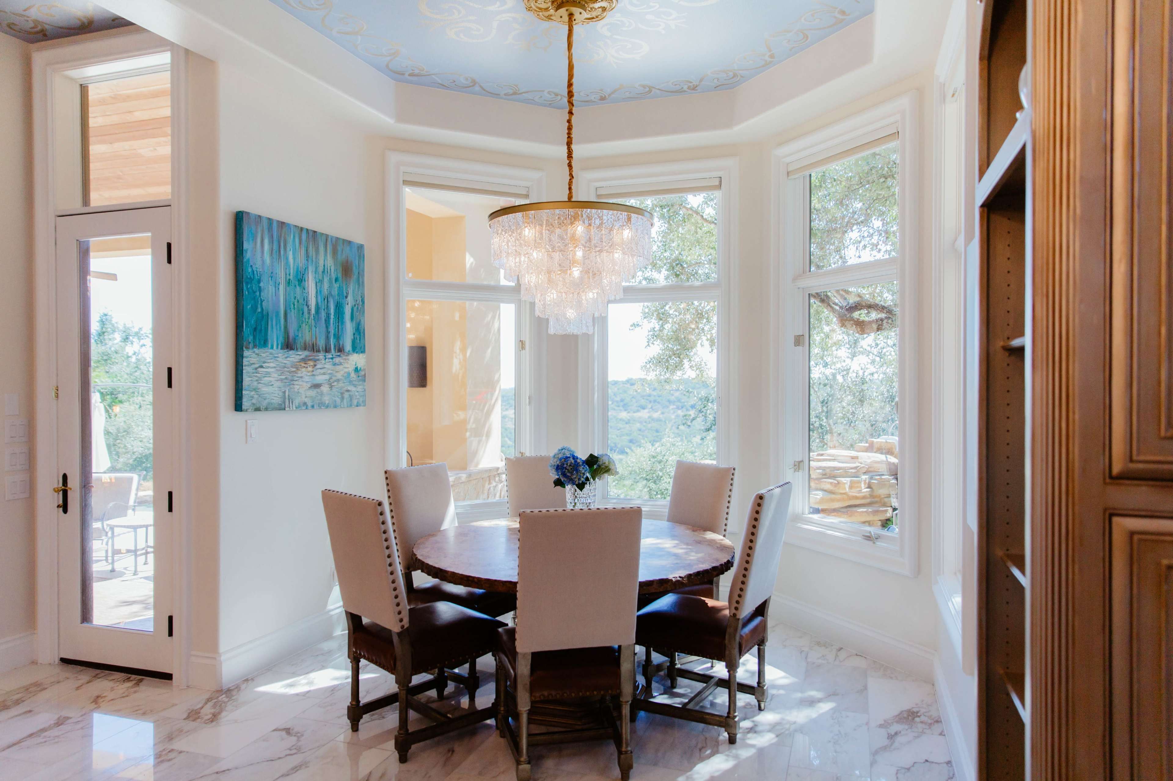 A round dining table with eight chairs is positioned under a chandelier in a room with large windows overlooking a view.