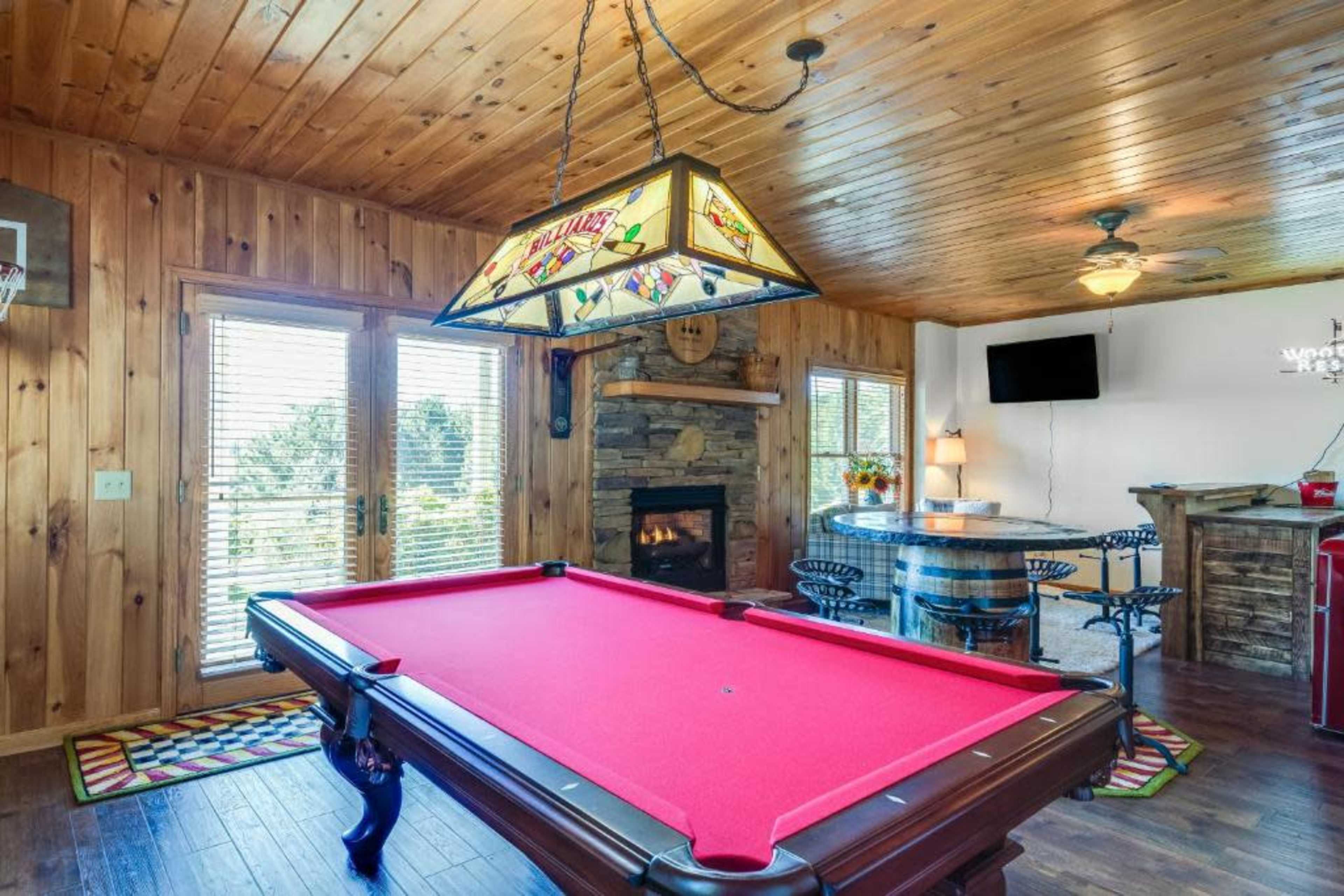 A recreational room with a red pool table, a stone fireplace, wooden walls, and a dining area featuring a round table and chairs.