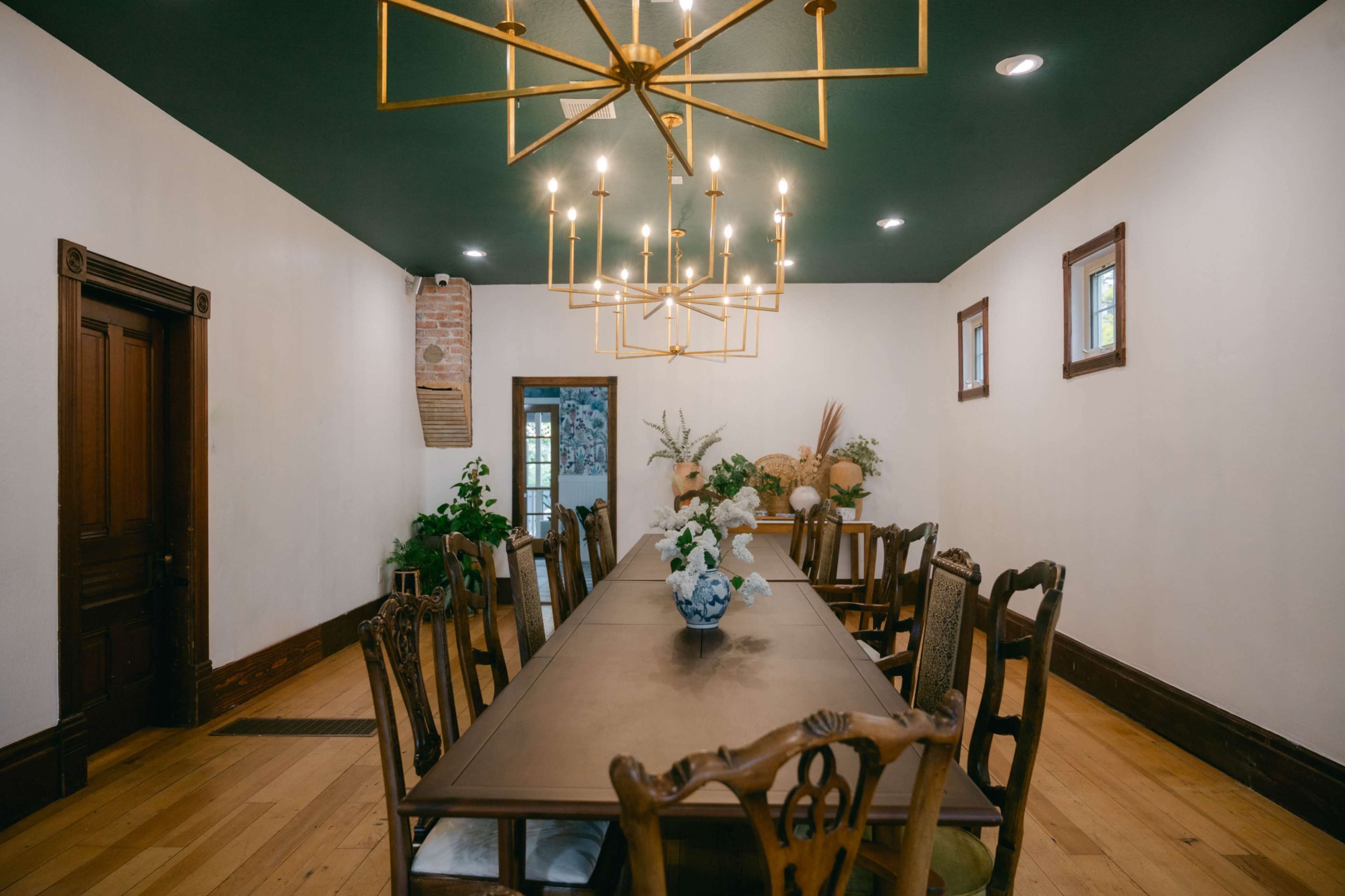 The image shows a spacious dining room with a long wooden table, surrounded by chairs, and a chandelier hanging from the ceiling.