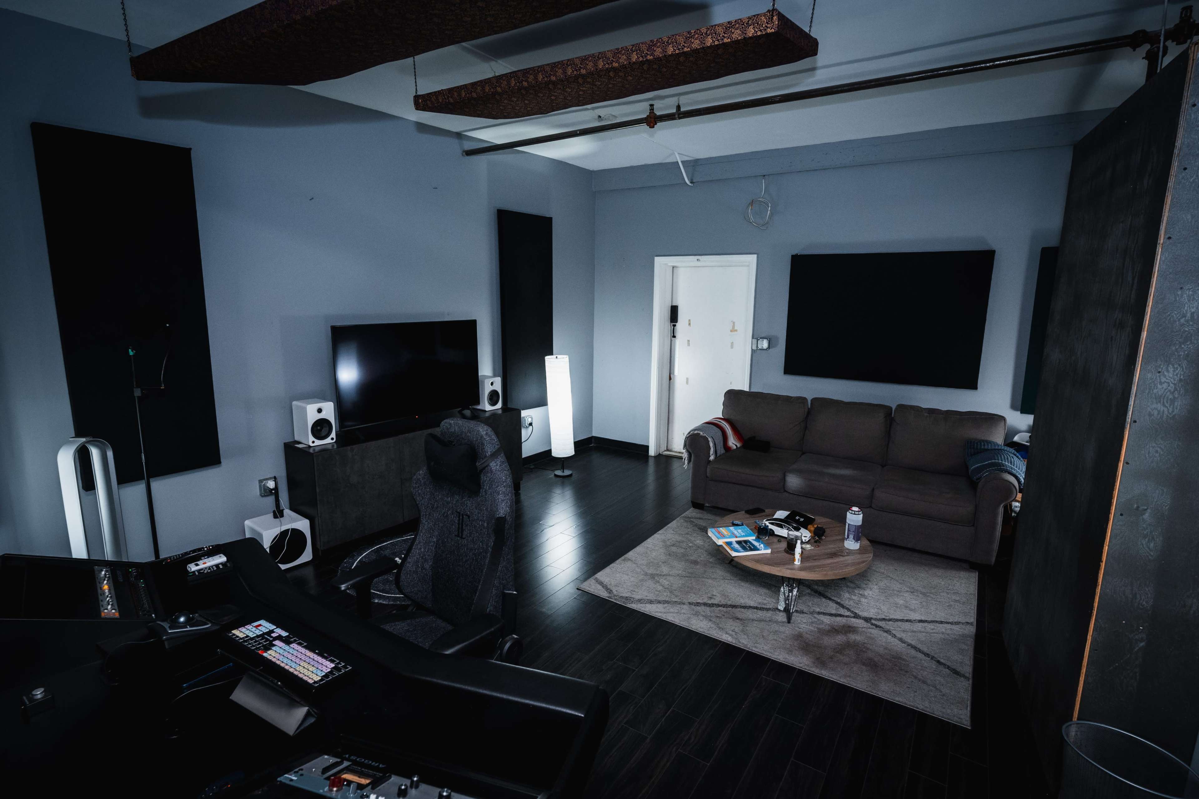 A music studio features a dark desk setup with audio equipment, a comfortable sofa, and flat-screen displays in a dimly lit room.