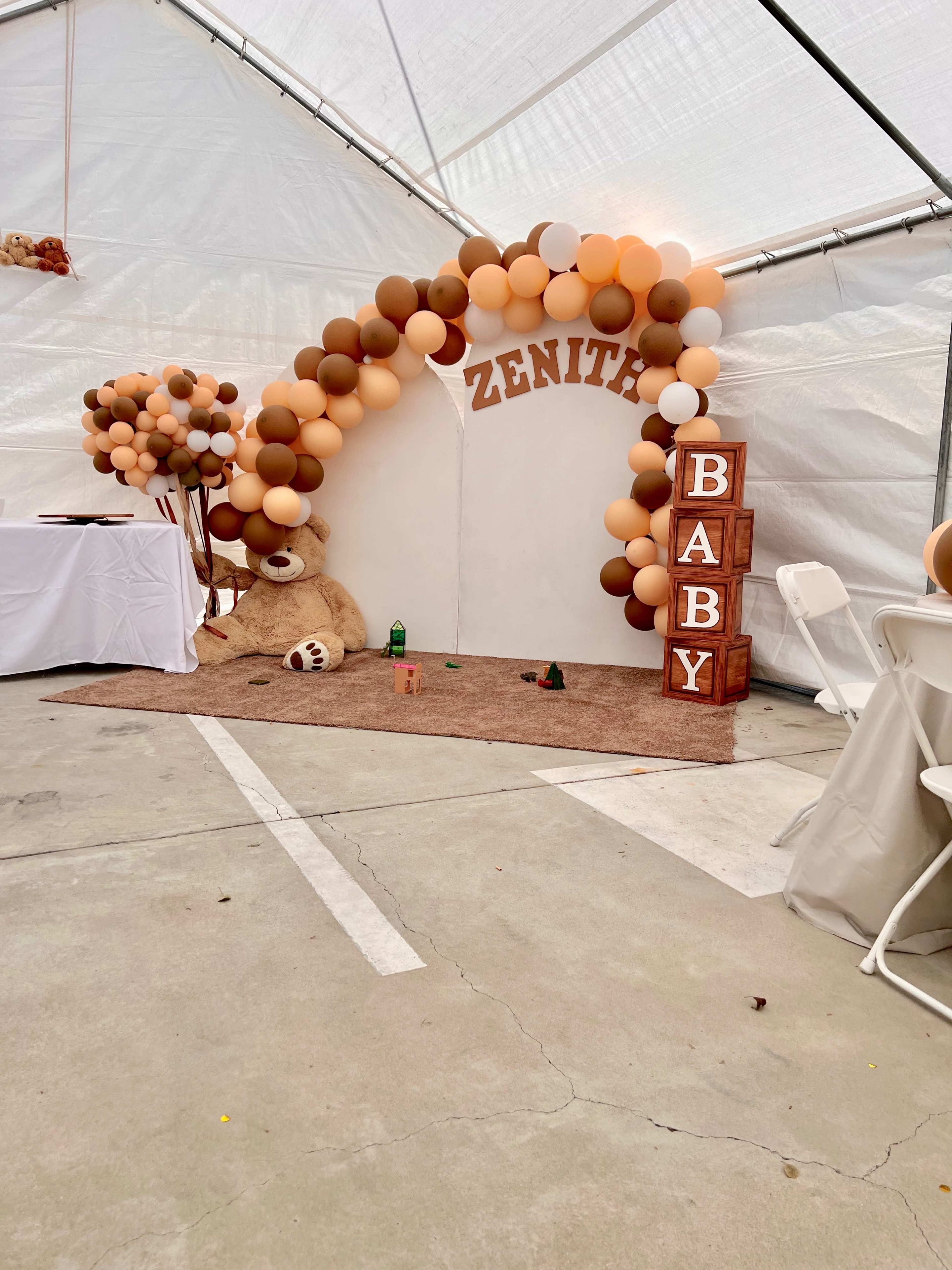 A party setup with a decorative arch made of brown and beige balloons, featuring the name "ZENITH" and a large teddy bear beside stacked wooden blocks that spell "BABY."