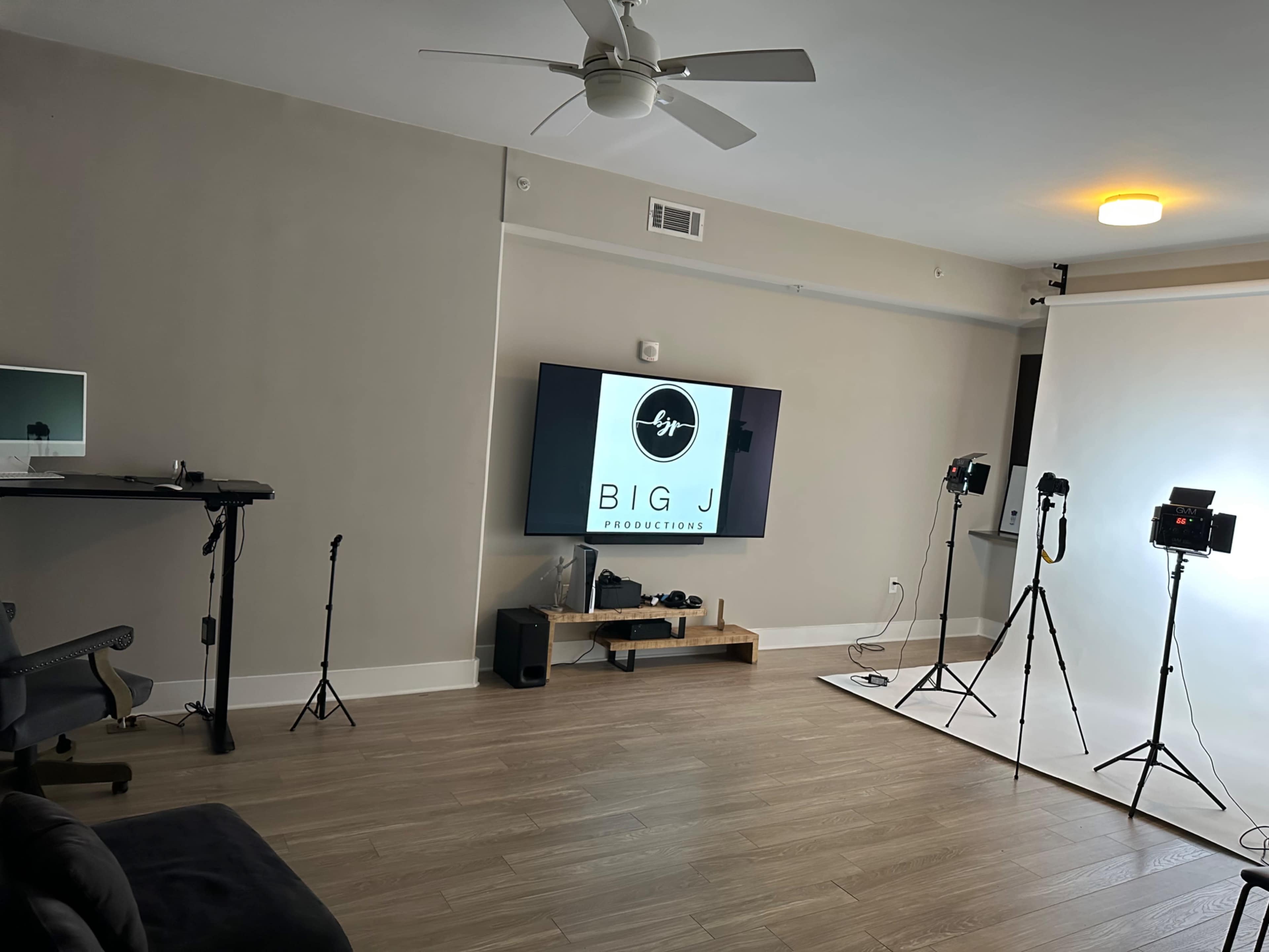 A minimalist studio space featuring a large wall-mounted screen displaying "BIG J PRODUCTIONS," along with lighting equipment and a backdrop for photography or video production.