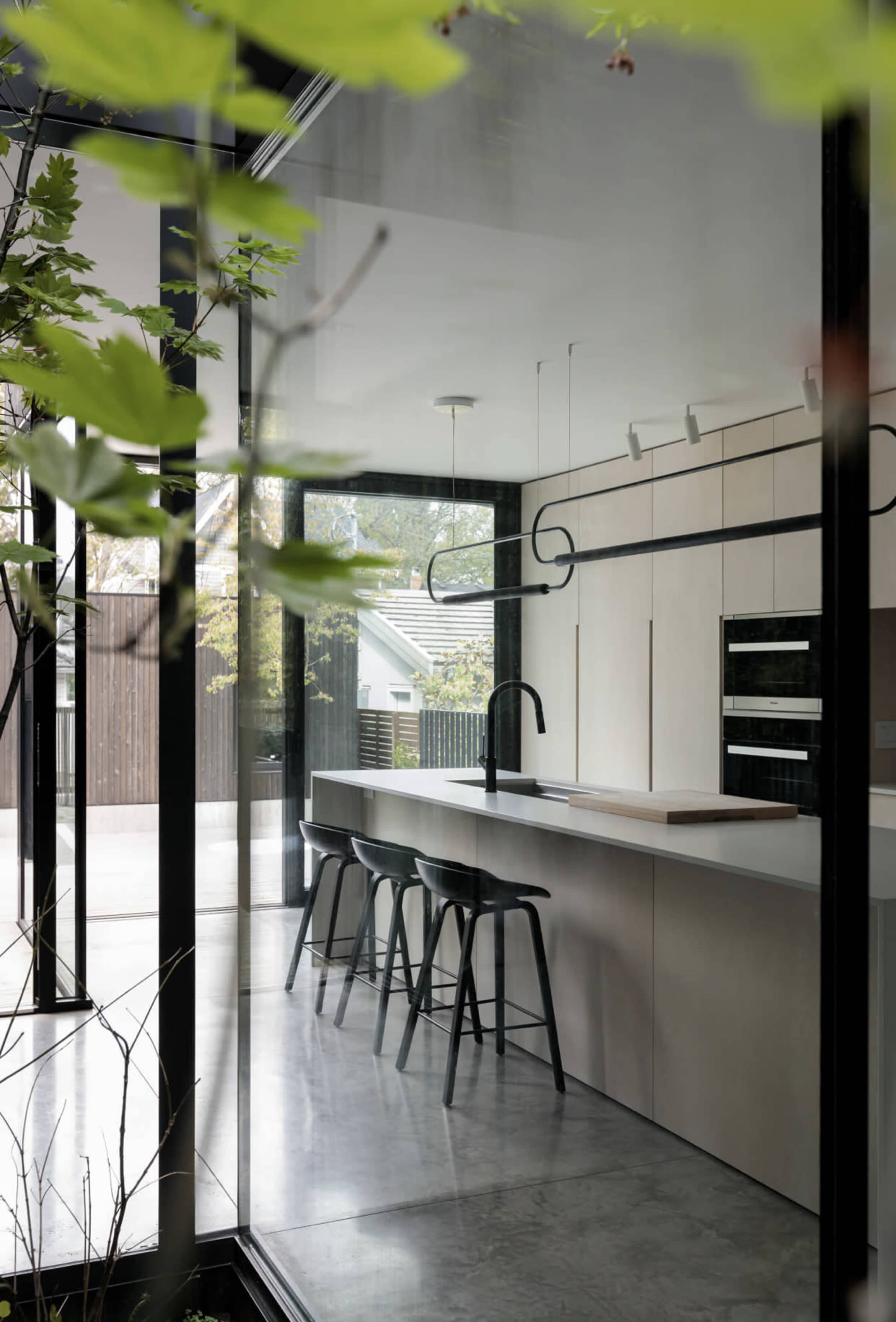A modern kitchen with a minimalist design, featuring a long countertop, black bar stools, and large glass walls that provide a view of the outdoor space.