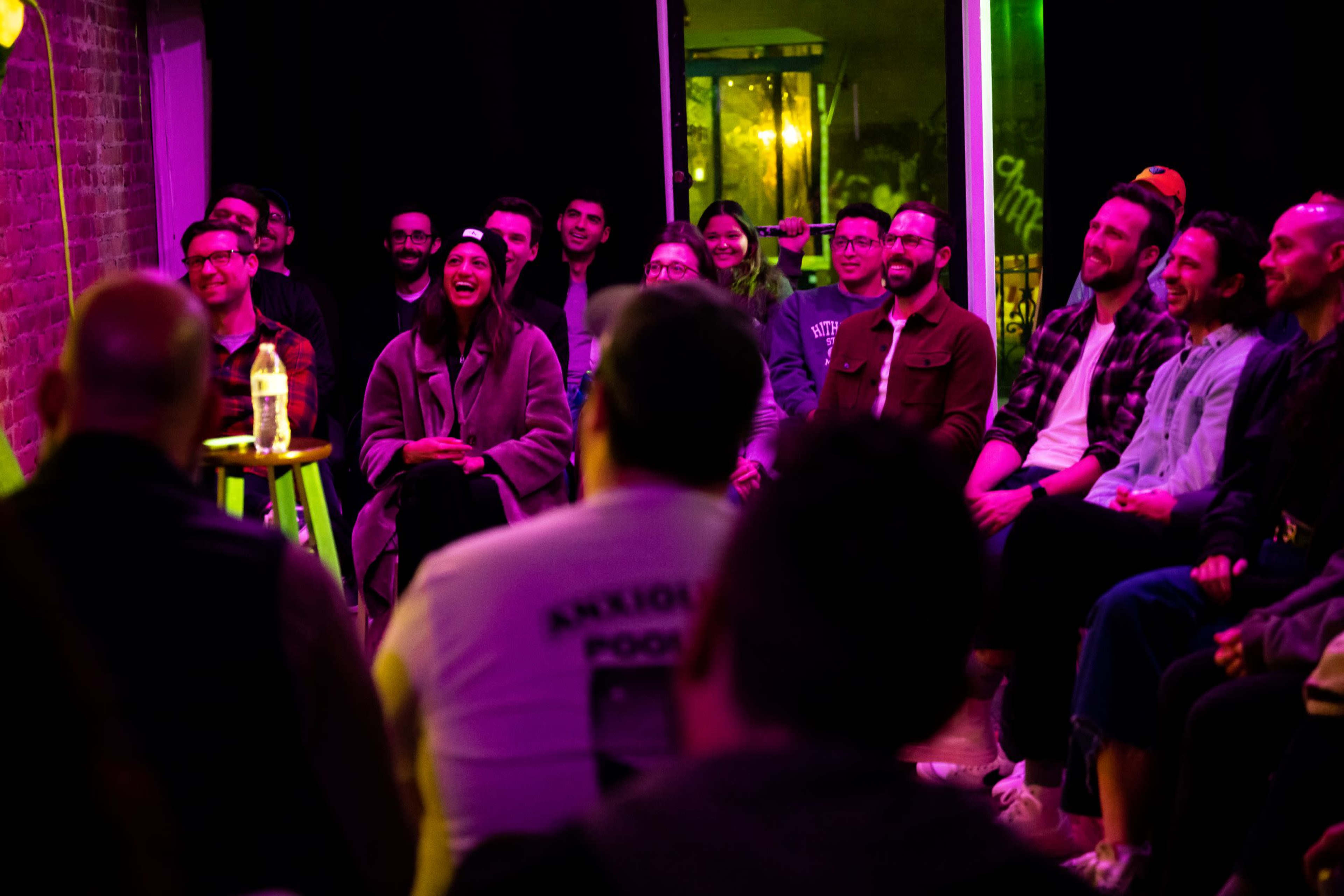 A group of people sits in rows, attentively watching a performer in a brightly lit venue.