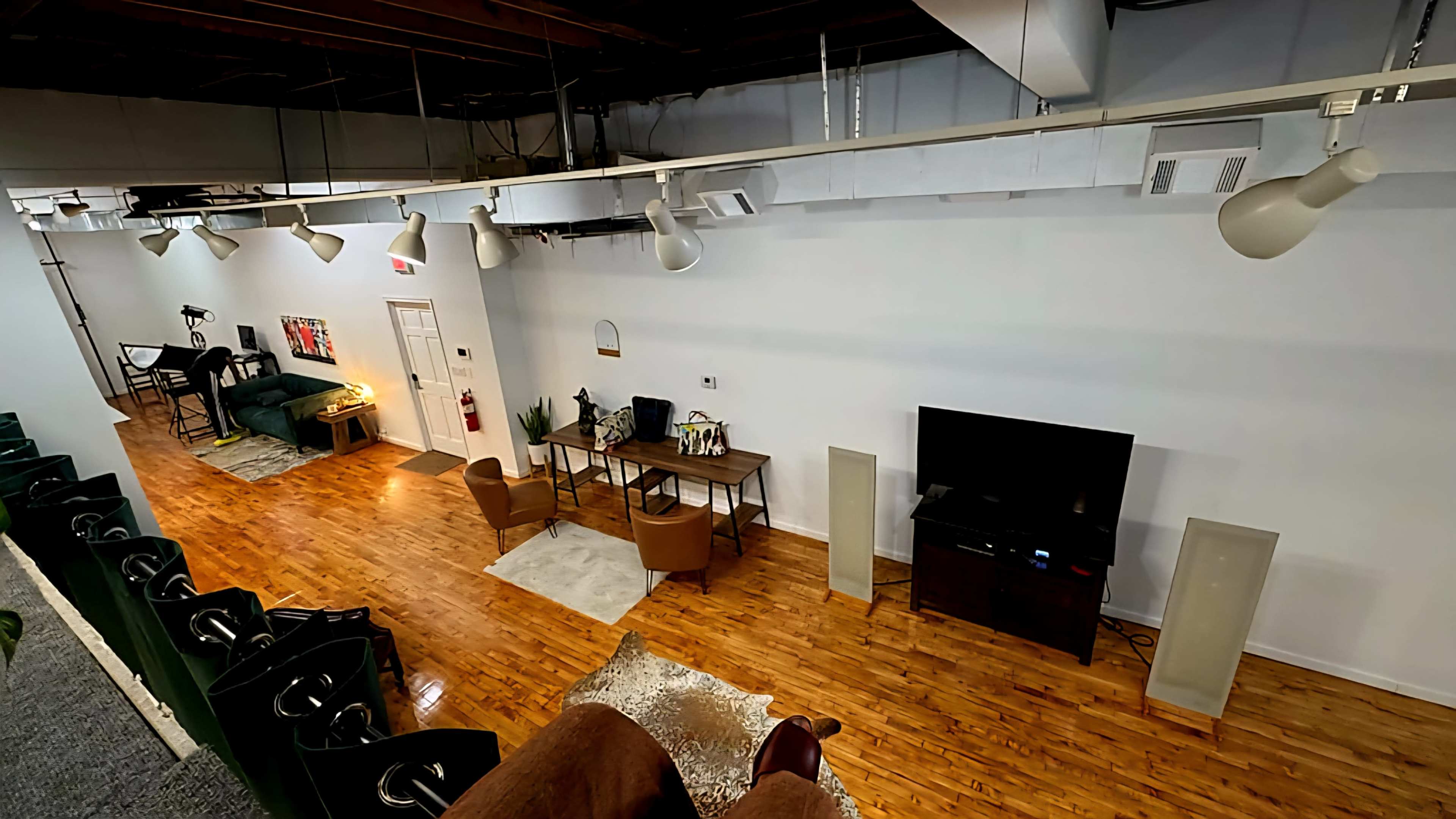 The image shows a spacious room with wooden flooring, featuring a seating area with two chairs, a desk, a television, and decorative elements along the walls.