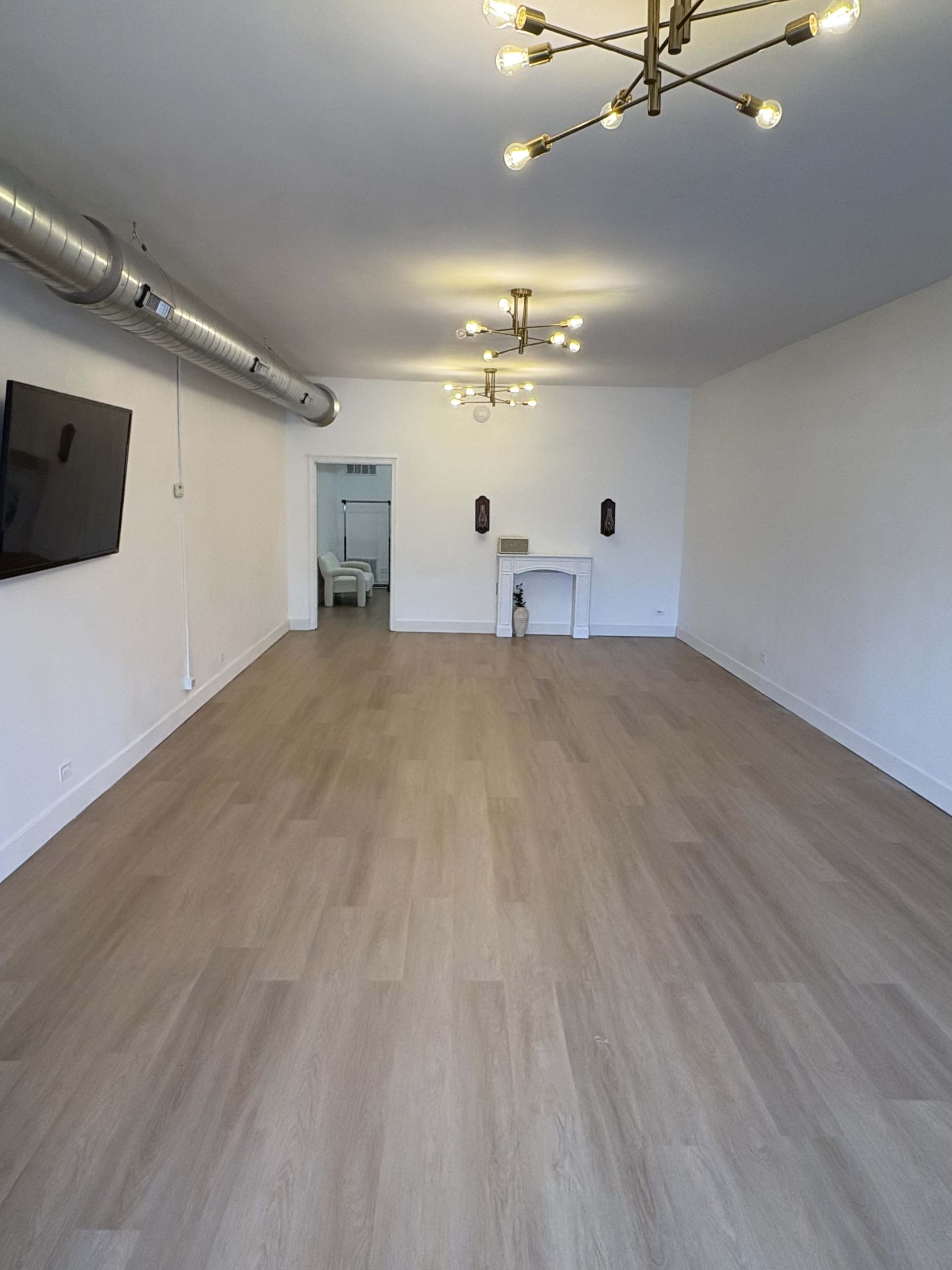 The image shows a spacious, empty room with light-colored wooden flooring and a modern ceiling light fixture, featuring a small fireplace against one wall.
