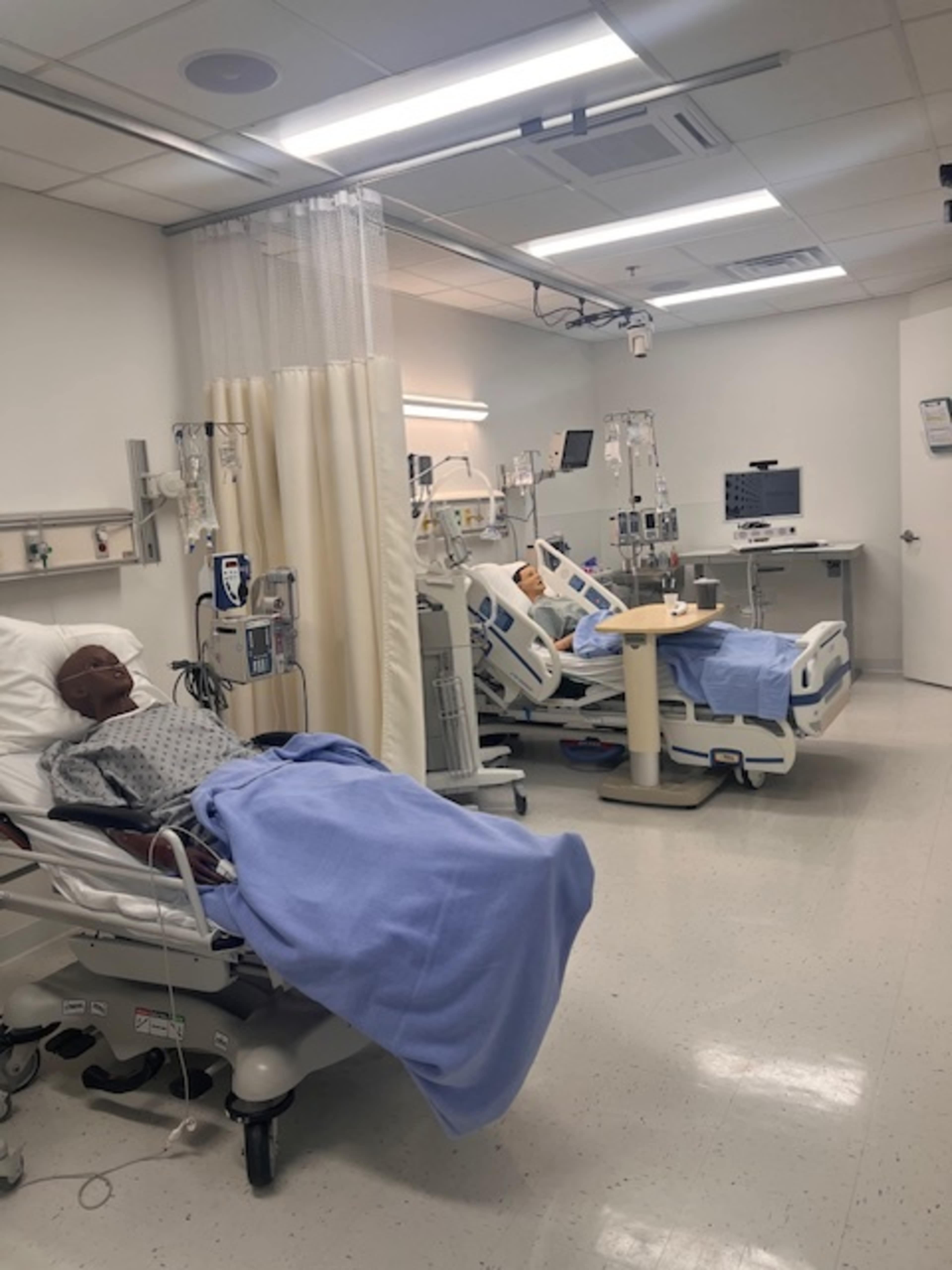 The image shows a hospital room with two beds containing medical mannequins, equipped with IV stands and monitors.